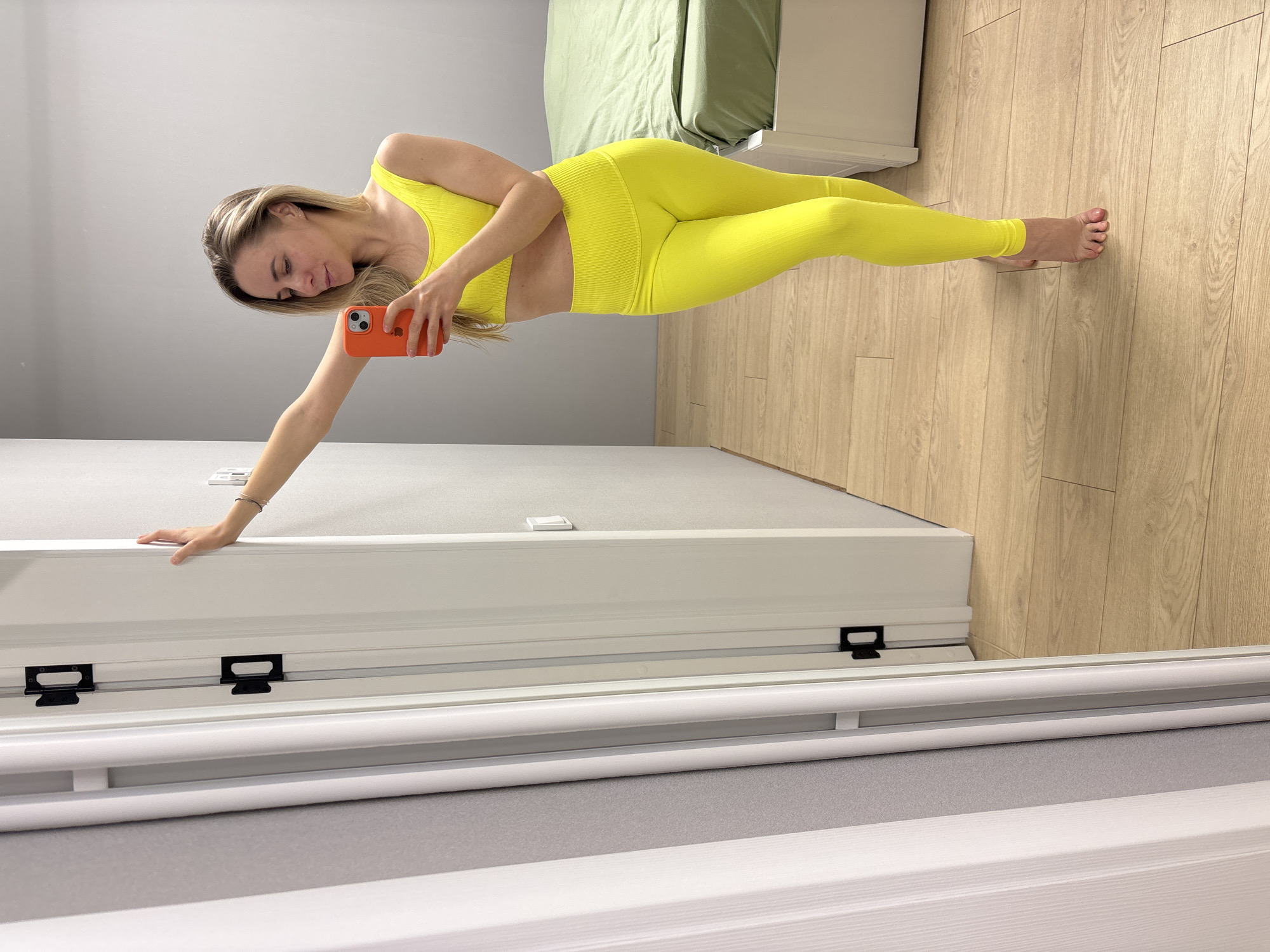 A blonde woman, appearing to be in her 20s or 30s, is captured in a mirror selfie inside what seems to be a bedroom or dressing area in Chaikovsky, Russia. She is wearing a vibrant, neon yellow ribbed two-piece athletic outfit consisting of a sports bra and high-waisted leggings. Her long, light-colored hair is styled down.
The woman is barefoot and holds an orange-cased smartphone, likely an iPhone, in her left hand, which is aimed towards the mirror. Her right arm is extended, with her hand resting on a white, flat surface, possibly the bottom frame of a large mirror or a piece of furniture, to steady herself while taking the photo. Her gaze is directed towards the phone screen as she poses.
The background of the image reveals a clean, modern interior. The floor is made of light-colored wooden laminate. The walls are painted a light grey. In the upper part of the reflection, a bed with a light green duvet or sheet is visible, suggesting a personal space. The mirror itself has a white frame along the bottom, with two black metal brackets attached, which could be part of a sliding door mechanism for a wardrobe. The lighting is bright and even, indicating daytime. There is no visible text in the image.
