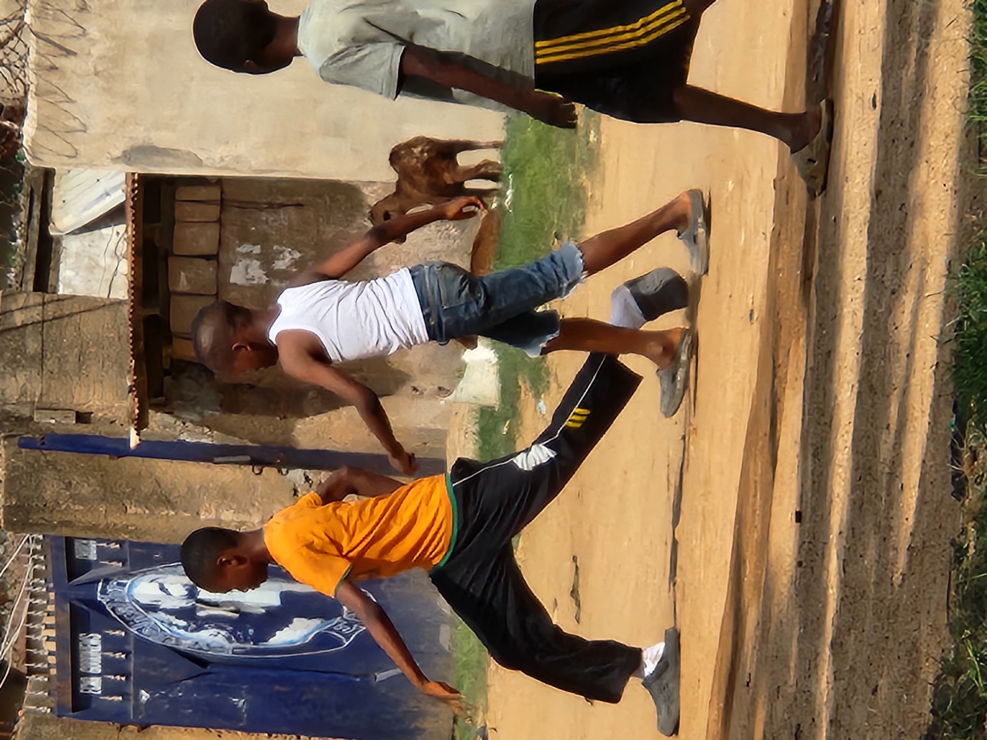 Three young boys are walking on a dirt path in Sabon Tasha, Nigeria. In the foreground, a boy wearing an orange t-shirt and black track pants with white and yellow stripes on the side walks towards the left. He is wearing grey Crocs-style sandals. To his right and slightly behind him, a second boy in a white tank top and blue denim shorts walks towards the right. A third boy, wearing a grey t-shirt and black shorts with yellow stripes, is further to the right, partially obscured by the other two, and is facing away from the camera. In the background, a brown calf stands on a grassy area near a light-colored building. To the left, a large blue gate with a circular emblem and white text is visible, set against a weathered wall. The environment appears dry and dusty, typical of a sun-drenched urban area.