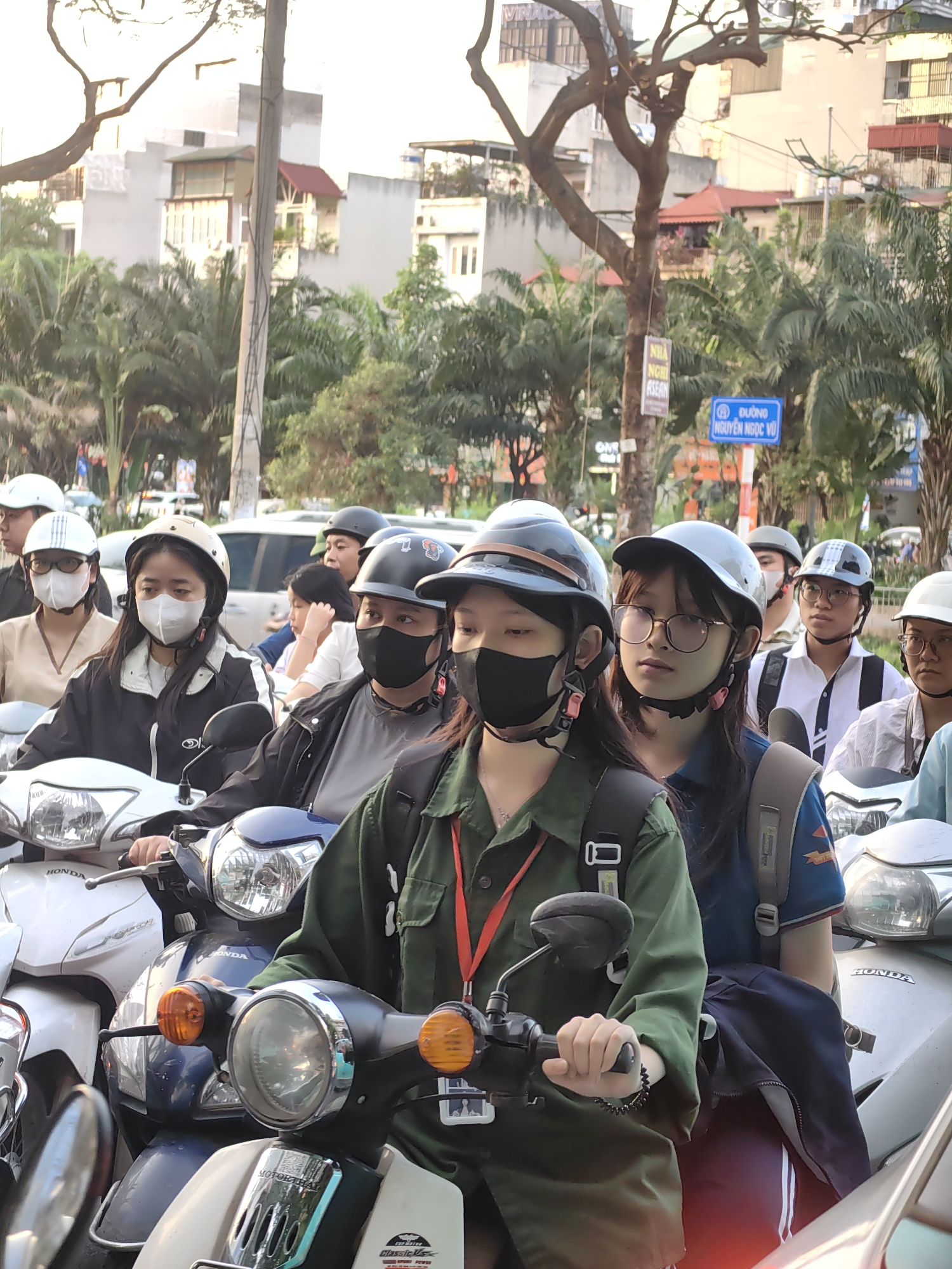 The image depicts a vibrant, urban street scene in Hà Nội, Vietnam, characterized by a large group of people on motorbikes, tightly packed as if in traffic or waiting at an intersection. The subjects are predominantly young adults and teenagers, many of whom are wearing protective helmets and face masks, a common sight in Vietnamese cities for safety and to guard against pollution.
In the foreground, a rider wearing a green utility-style jacket and a black mask grips the handlebars of a scooter, with a passenger behind her in a blue shirt and glasses, carrying a backpack. To their left, another person in a black and white jacket and a white mask rides a white Honda scooter, while a passenger behind them wears a beige top and glasses. Numerous other individuals on scooters with various helmet designs and masks fill the midground, indicating a busy thoroughfare.
The scene is set against a backdrop of multi-story buildings with different architectural styles and abundant palm trees, suggesting a tropical or subtropical climate. The lighting appears soft and warm, characteristic of daytime, possibly late afternoon.
Visible text provides strong location context. A blue road sign clearly states "ĐƯỜNG NGUYỄN NGỌC VŨ," a street in Hà Nội. Another sign on a tree reads "NHÀ NGHỈ ASEAN," identifying an "ASEAN Guesthouse." Brand names like "HONDA" are visible on several scooters, along with "MOTORTHAI," "Classic V...", and "SPUR POWER." A partially visible sign on a distant building reads "VINACON." The overall image conveys the energetic atmosphere of a typical day in a bustling Southeast Asian city.