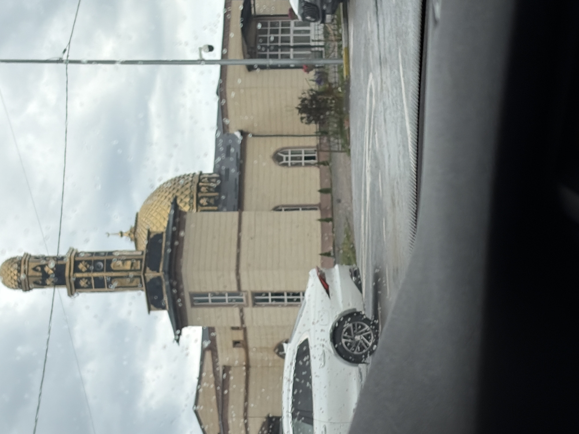 An overcast, rainy day is depicted from the perspective of someone inside a vehicle. Water droplets are visible on the car's windshield, obscuring the view of a building and its surroundings.
The building, appearing to be a mosque, features a golden dome and a tall minaret adorned with gold and black accents. The mosque's facade is a pale yellow, with rectangular windows and arched windows. A white car is parked in front of the mosque, and its wheels are visible. The ground is wet, indicating recent rainfall. A lamppost with a security camera is also present. The image is a medium shot with the foreground dominated by the interior of a car, likely the dashboard. The scene is calm and uneventful.