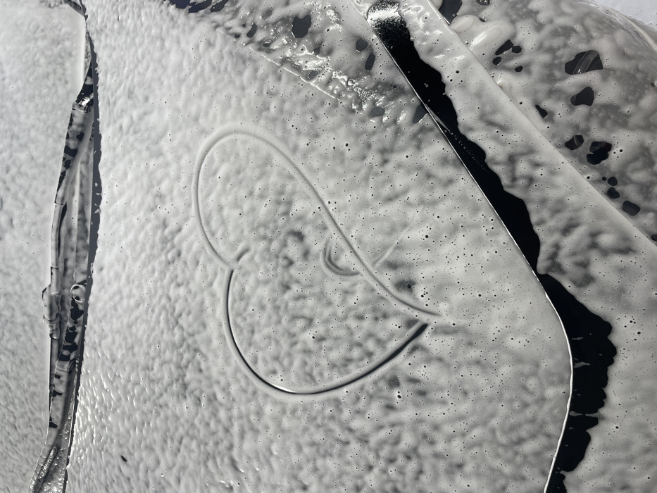 The image presents a close-up, top-down perspective of a vehicle's hood and lower windshield, densely coated in white car wash foam. A prominent heart shape has been drawn into the foam on the vehicle's hood, revealing the dark surface underneath. Within the larger heart, a smaller, incomplete swirl is discernible. The base of the windshield is visible at the top of the frame, with two dark windshield wipers positioned horizontally and partially covered in foam. The vehicle's paint is a dark color, visible where the foam is disturbed or thin. No individuals are depicted. The scene is located in Chaikovsky, Russia.