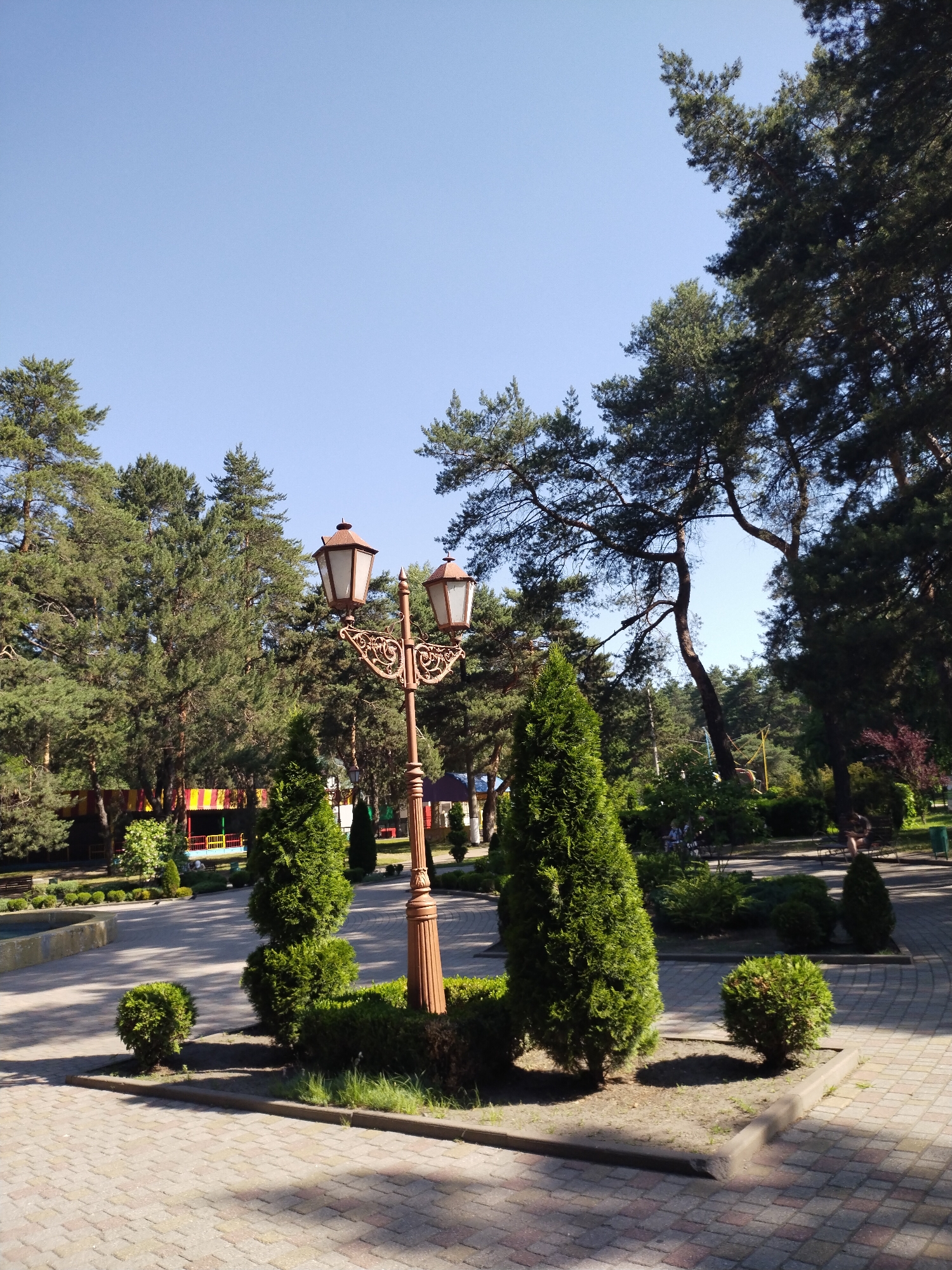 The image depicts a park scene in Horishni Plavni, Ukraine. A decorative lamppost with two lights stands prominently in the foreground, casting shadows on a paved walkway. The walkway is made of light-colored bricks arranged in a patterned fashion. Surrounding the lamppost are manicured bushes and two evergreen trees, one cone-shaped and another spherical. In the background, a line of tall pine trees borders the park, with a clear blue sky visible above. Further back, hints of recreational facilities, including colorful structures with yellow, red, and blue elements, suggest a playground or amusement area. Several park benches are visible, some occupied by individuals, though their faces are not discernible. The overall environment is one of a well-maintained public park on a sunny day.