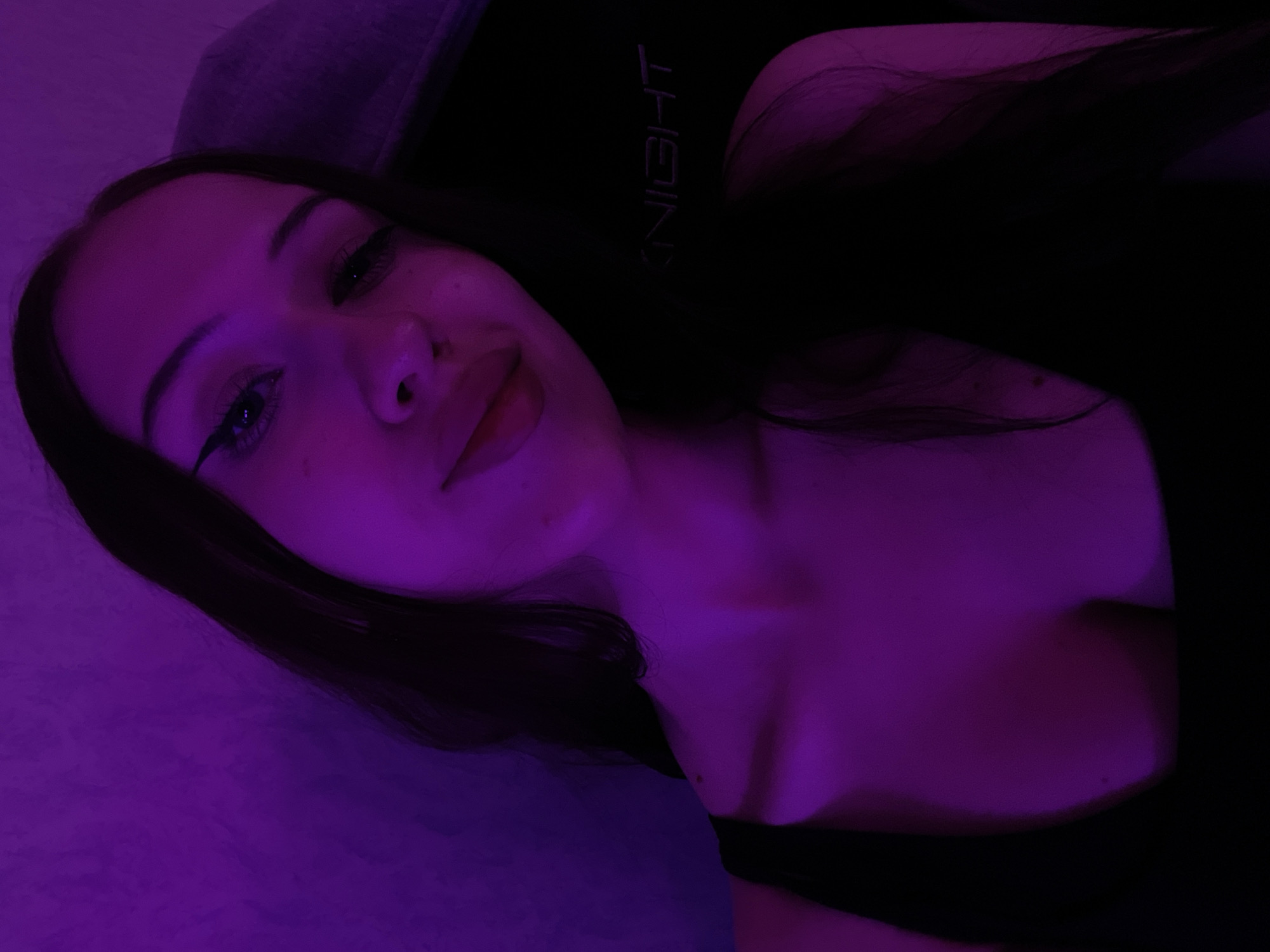 A young woman with long, dark hair is captured in a selfie. She is wearing a black top and has noticeable eyeliner. The lighting is predominantly purple, casting a distinct hue over the scene and her skin. The background appears to be a textured wall. Behind her, to the right, a dark object with the word "KNIGHT" visible is present, possibly a piece of furniture or equipment. The image does not provide any information regarding a specific location in the city of Votkinsk, Russia.