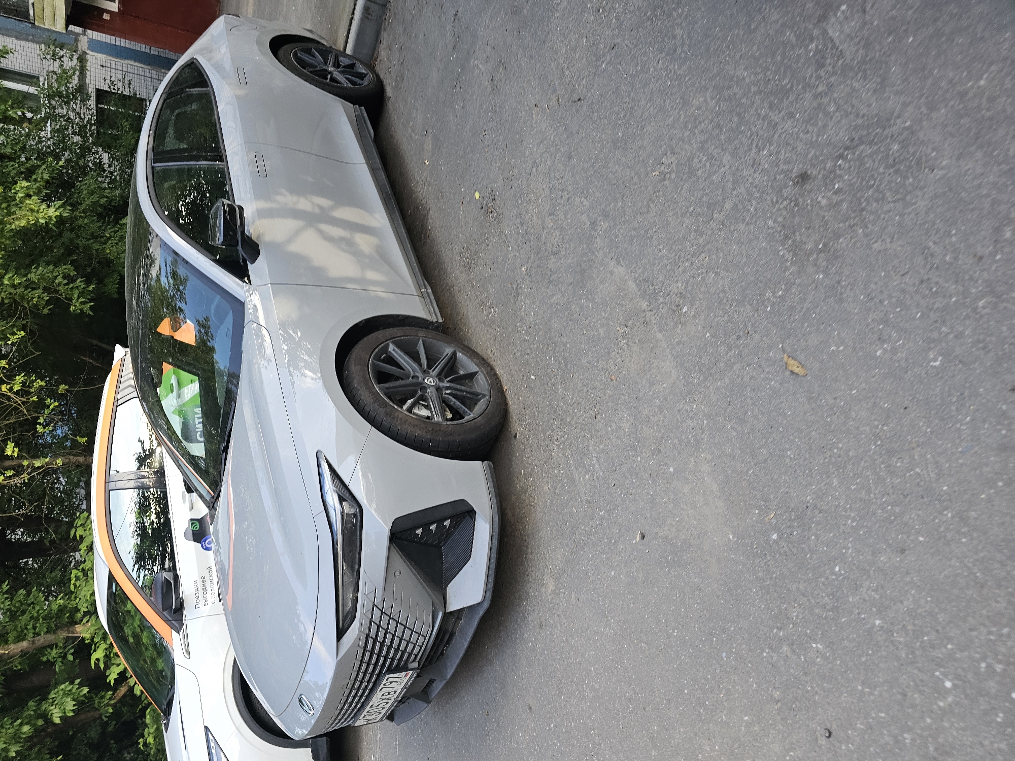 Two cars are parked on an asphalt surface in front of a building and trees. The car in the foreground is a light gray sedan with a black roof and black rims. Its license plate reads "X305XB 797". The car in the background is a white SUV with an orange accent stripe on the roof and sides. The SUV has text on its door that reads "Поездки выходного дня с Подпиской".