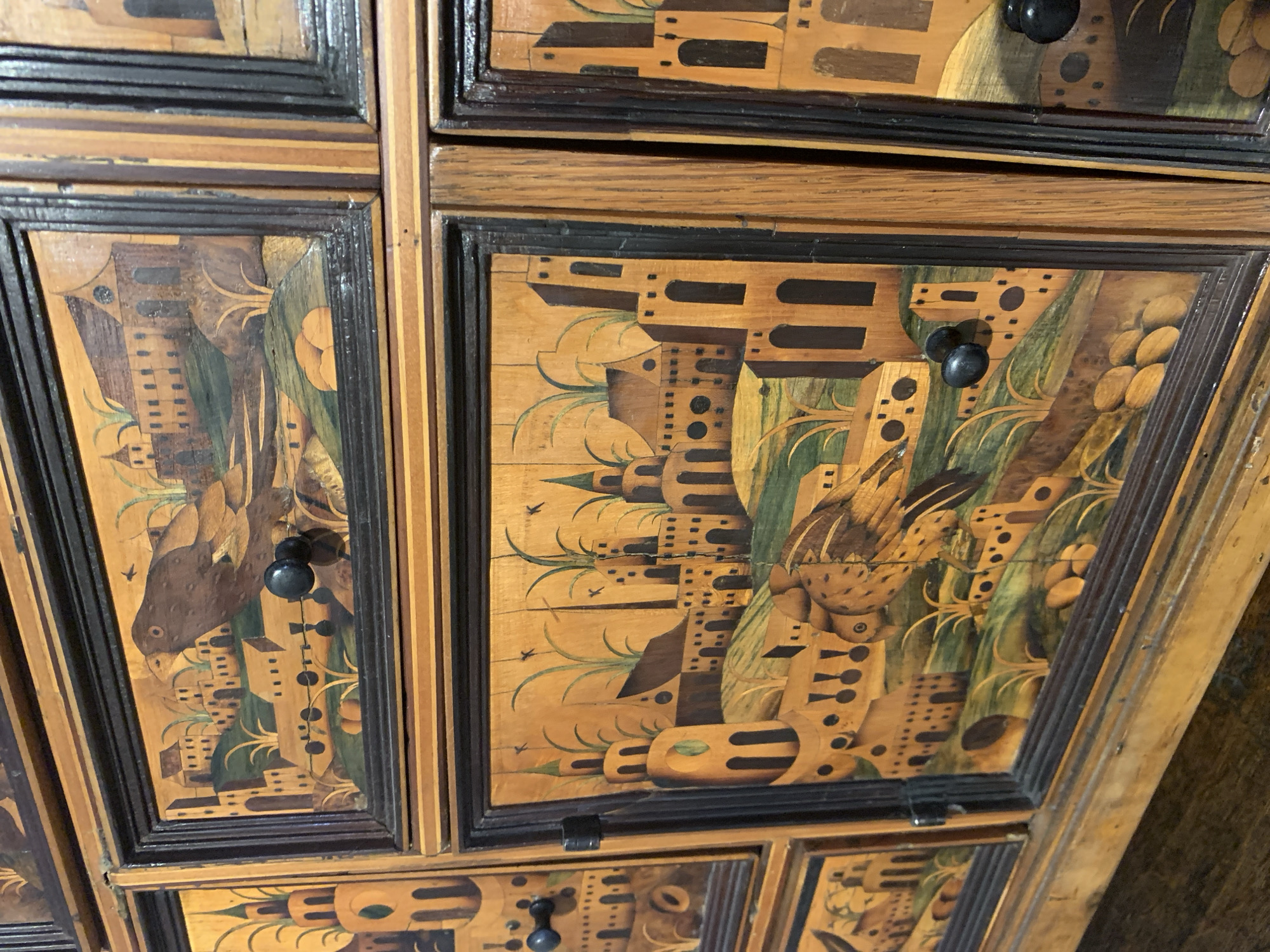 The image displays a close-up view of intricately decorated antique wooden drawers or cabinets. The decorative elements are inlays of various wood types, creating scenes of birds and stylized buildings. The artwork is detailed and colorful, with a mix of warm wood tones, greens, and browns. Some drawers have black circular knobs. The scene is set in the city of Poznan, Poland.