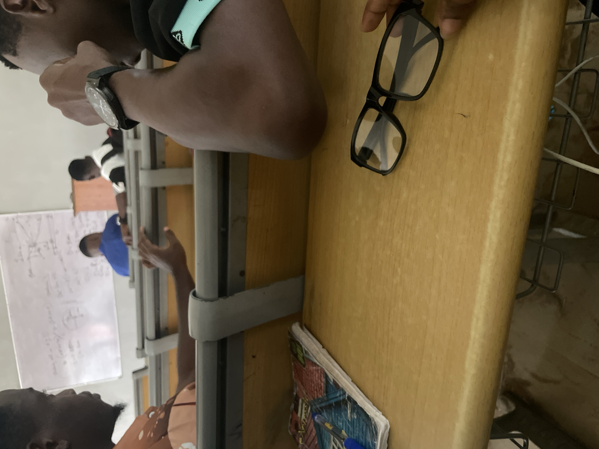 The image depicts a classroom setting, possibly a lecture hall, in Ihiagwa, Nigeria. In the foreground, a wooden desk holds a pair of black-framed glasses and a notebook with a blue pen resting on it. The notebook's cover has a colorful, possibly comic-book or abstract design. A person's hand is visible reaching for the glasses. In the background, a whiteboard displays handwritten equations and diagrams, indicative of an academic lesson. Several students are seated at tiered desks. One student in the mid-ground wears a blue shirt, and another in the far background wears a white shirt with black stripes. The overall environment suggests an educational institution.