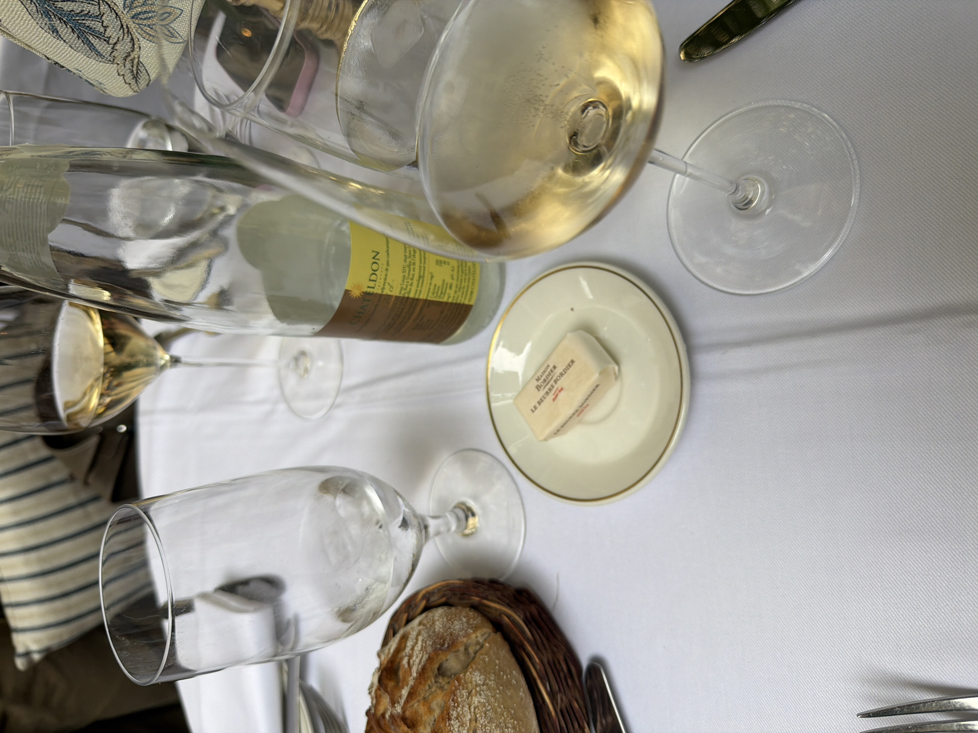 The image depicts a table setting, likely in a restaurant in Paris, France. A bottle of CHATELDON sparkling water is lying on its side, near a full wine glass. Next to it, there is a small plate with a square of butter from "Maison Beurre Bordier". Further down, a basket of bread is placed beside an empty wine glass. Cutlery is visible at the bottom of the frame. A patterned napkin partially appears at the top-left corner. The tablecloth is plain white.