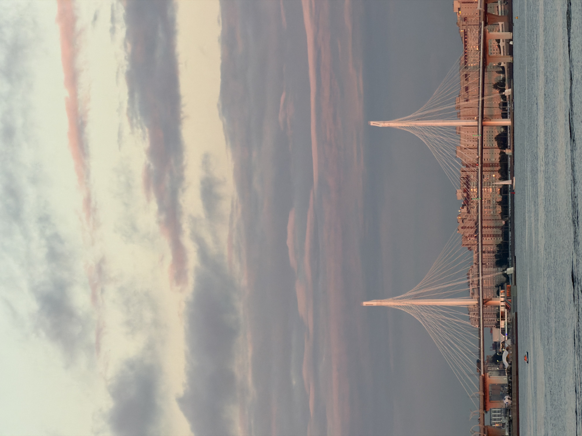 The media file depicts a cable-stayed bridge spanning a body of water, likely a river or bay, adjacent to a city. The sky is filled with dramatic clouds, tinged with pink and orange hues, suggesting either sunrise or sunset. The lighting is soft, indicating it's either early morning or late evening. The cityscape features multi-story buildings that are illuminated, adding a warm glow against the twilight sky. The water is calm with gentle ripples. Two cable towers of the bridge are prominently visible, supporting a network of tensioned cables that extend to the bridge deck. The bridge appears to be modern and elegantly designed. In the water, a small boat can be seen, adding a sense of scale. There is no visible text in the image. The overall mood is serene and picturesque, capturing the beauty of the urban landscape at dusk or dawn.