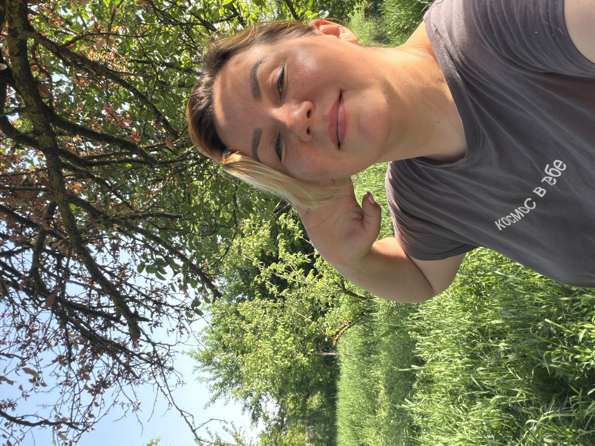 A woman is taking a selfie outdoors. The background shows a field of tall grass and green trees under a bright sky with tree branches and leaves partially obscuring the view. The woman has light skin, fair hair, and is wearing a gray t-shirt with Cyrillic text on it. The location is Leningradskaya, Russia, based on the text on the t-shirt.