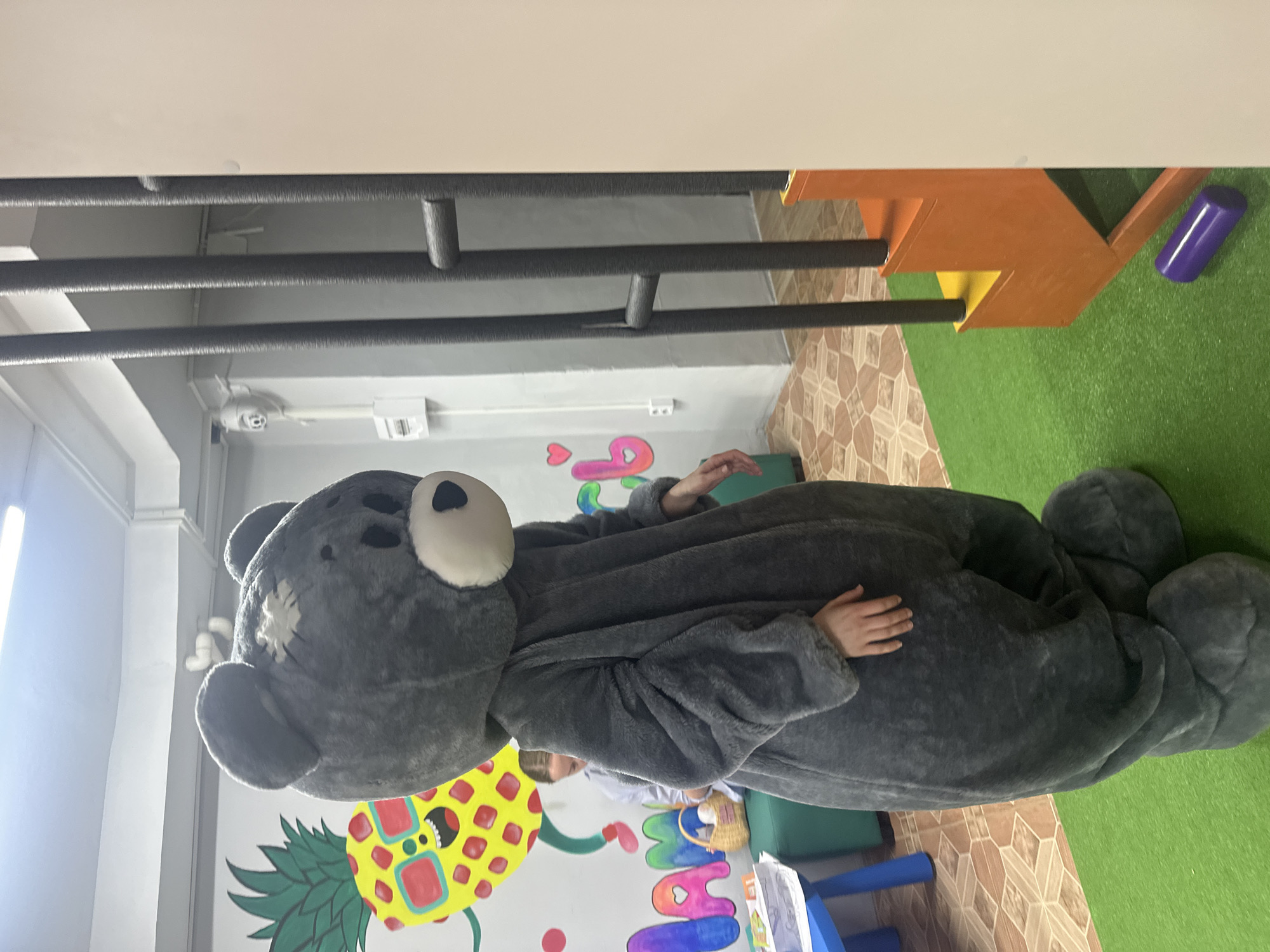 The image shows an indoor scene, likely a children's play area or event venue. A person is prominently featured, wearing a large, plush, dark grey bear costume. The bear costume has a white muzzle, black nose, and black eyes, with a distinctive white stitched patch on the left side of its head. The wearer's bare hands are visible emerging from the costume's sleeves, with one hand resting on the bear's midsection and the other slightly raised.
Behind the person in the costume, a child is partially visible, looking downwards, next to a small woven basket. The floor in the room is split, with green artificial turf on the right side and brown patterned tiles on the left. The white walls are decorated with colorful murals, including a large yellow pineapple illustration with a face and several brightly colored, stylized letters. The visible letters spell out "HAPP," which appears to be part of a larger word like "HAPPY."
In the upper portion of the image, multiple black padded bars or pipes run horizontally, possibly serving as protective elements or part of a play structure. On the right side of the frame, there's an orange and yellow play structure, and a purple cylindrical object lies on the green turf. The setting, with its playful decor and child-sized elements, suggests a festive or entertainment-focused environment, with the person in the bear costume likely serving as an entertainer or mascot.