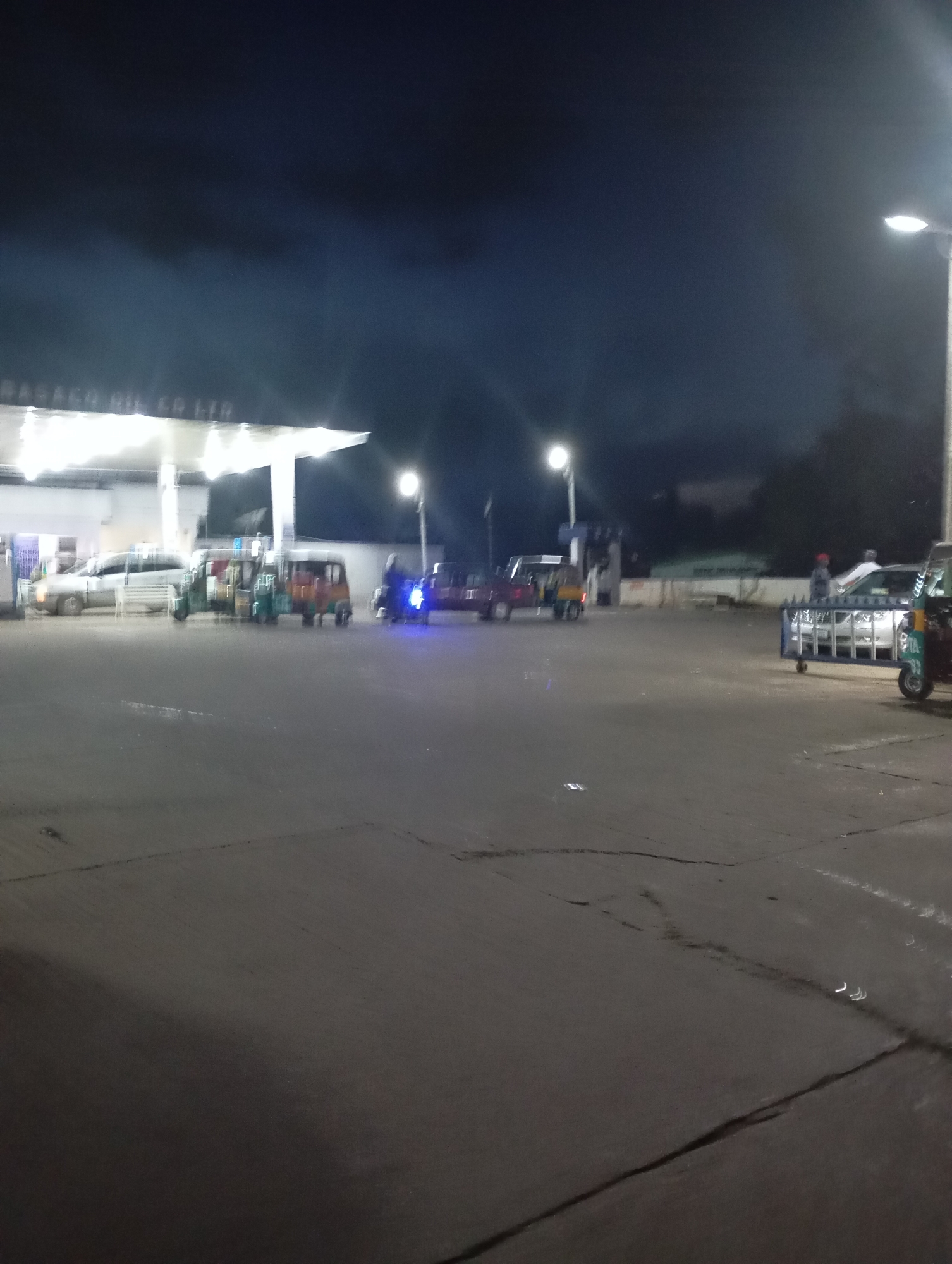 The image captures a nighttime scene at a gas station in Jos, Nigeria. A white canopy with illuminated signs, likely indicating the gas station's name, is visible in the background. Several vehicles are present, including what appear to be auto-rickshaws, also known as "Keke NAPEPs," and a van. One auto-rickshaw is parked in the foreground on the right, its front illuminated by a blue light. Another auto-rickshaw is visible to the left, with a person seated in it. A white van is parked further back on the left. The ground is paved, and streetlights illuminate the area. The sky is dark, suggesting nighttime, with some clouds visible. The overall impression is that of a bustling gas station operating at night in Jos, Nigeria, with various modes of transportation present.