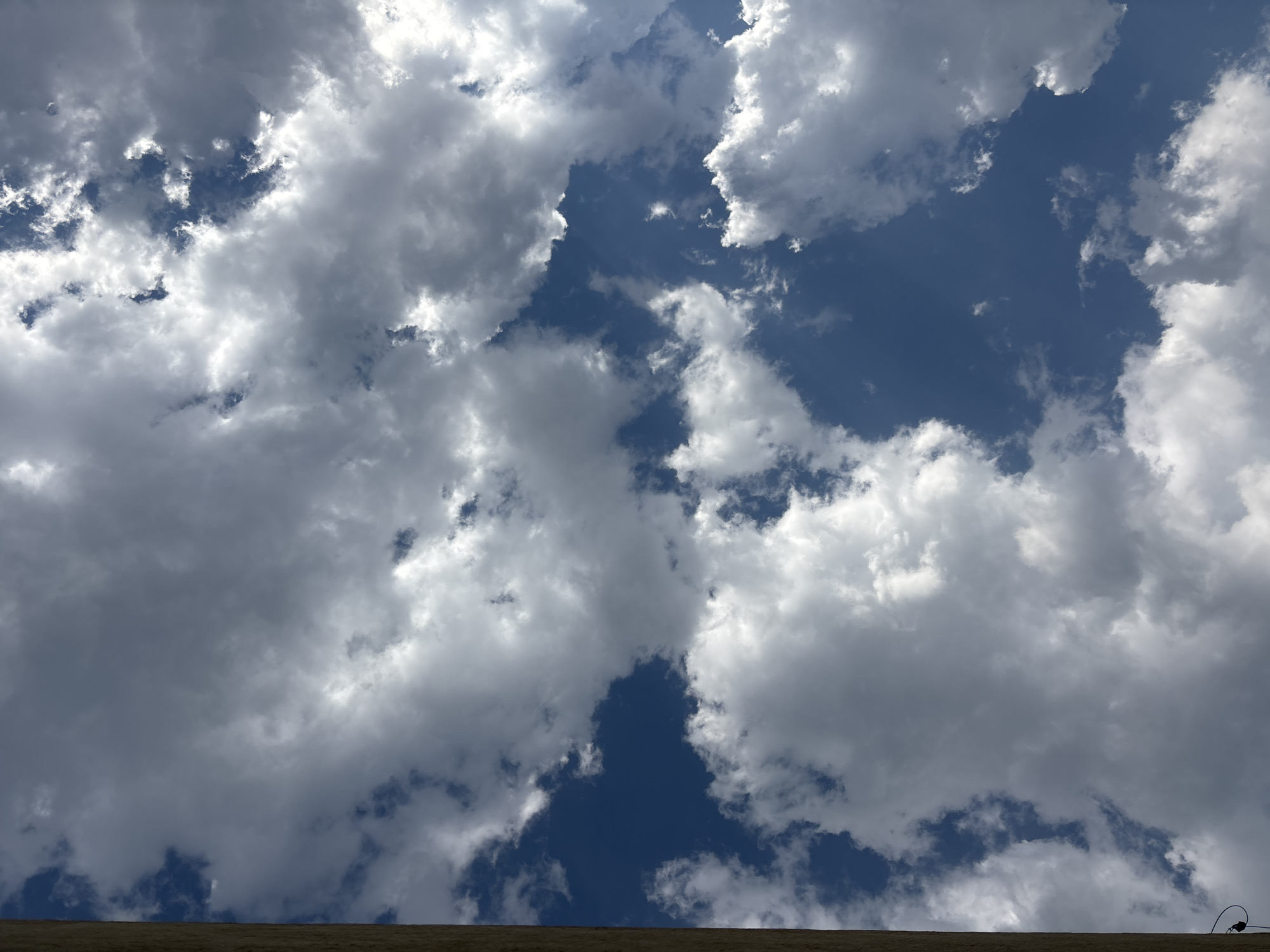 The image shows a view of the sky with numerous fluffy, white clouds against a blue backdrop. The perspective is looking upward, with a small portion of a brown, textured surface visible at the very bottom of the frame, possibly a rooftop or the edge of a building. In the lower right corner, a thin, dark wire curves into the frame from the edge of the building. The photograph appears to be taken outdoors during daylight hours. The location is Puebla, Mexico.