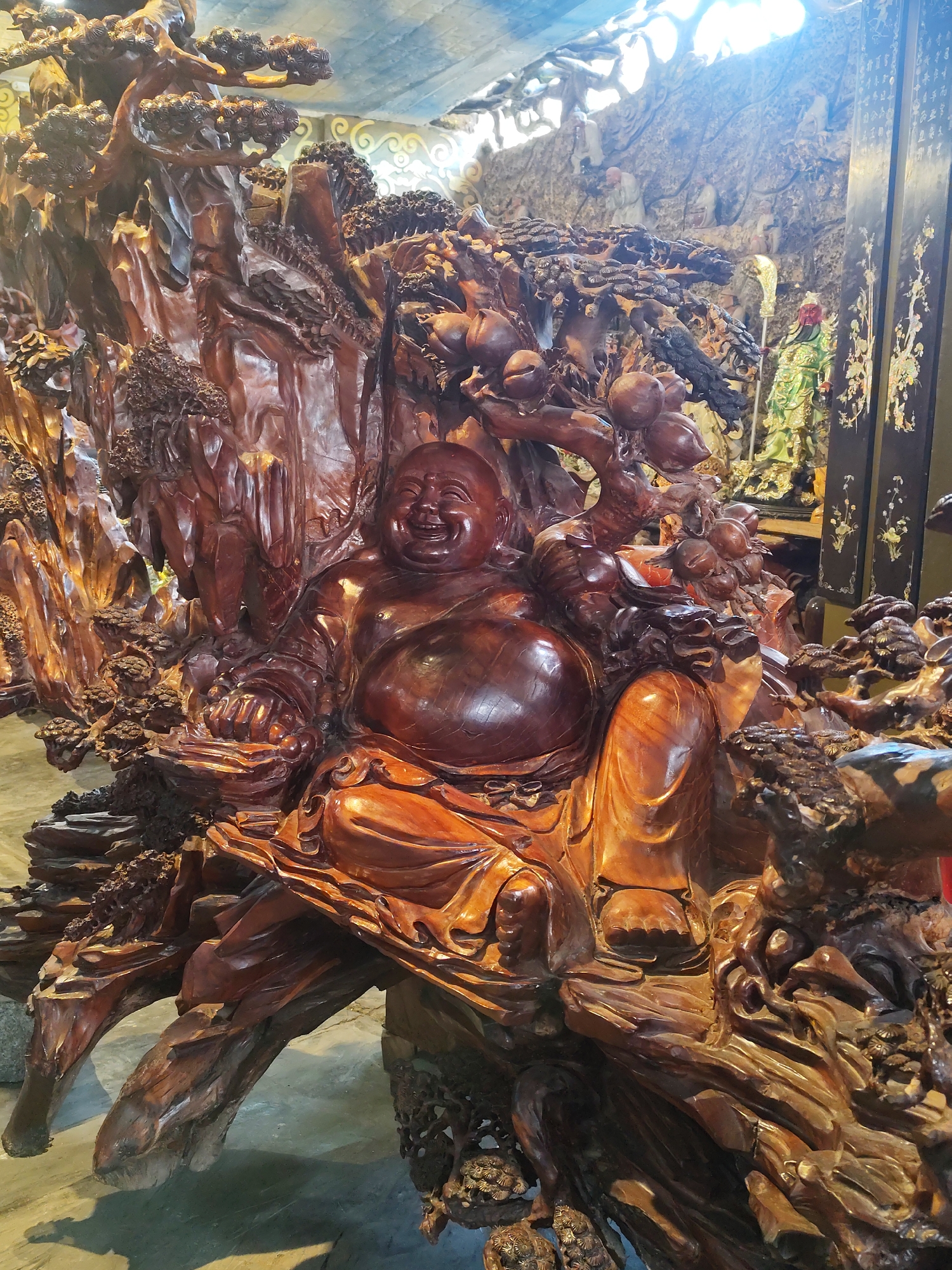 A large, intricately carved wooden sculpture depicts a laughing Buddha seated amidst a rocky, tree-covered landscape. The Buddha has a prominent belly and is dressed in flowing robes, with his legs crossed. His face is open in a wide, joyous smile, and his eyes are closed. The sculpture is made of dark, polished wood, with rich variations in color and texture. Surrounding the Buddha are detailed carvings of gnarled trees, rocks, and what appear to be fruit, possibly peaches. In the background, partially visible, is a mural or wall carving with figures and inscriptions, and a statue of a figure in a green and gold garment holding a weapon. The lighting suggests an indoor setting, possibly a temple or a place of worship, with a bright light source coming from above and behind the sculpture, casting shadows. The overall impression is one of opulence and spiritual significance.