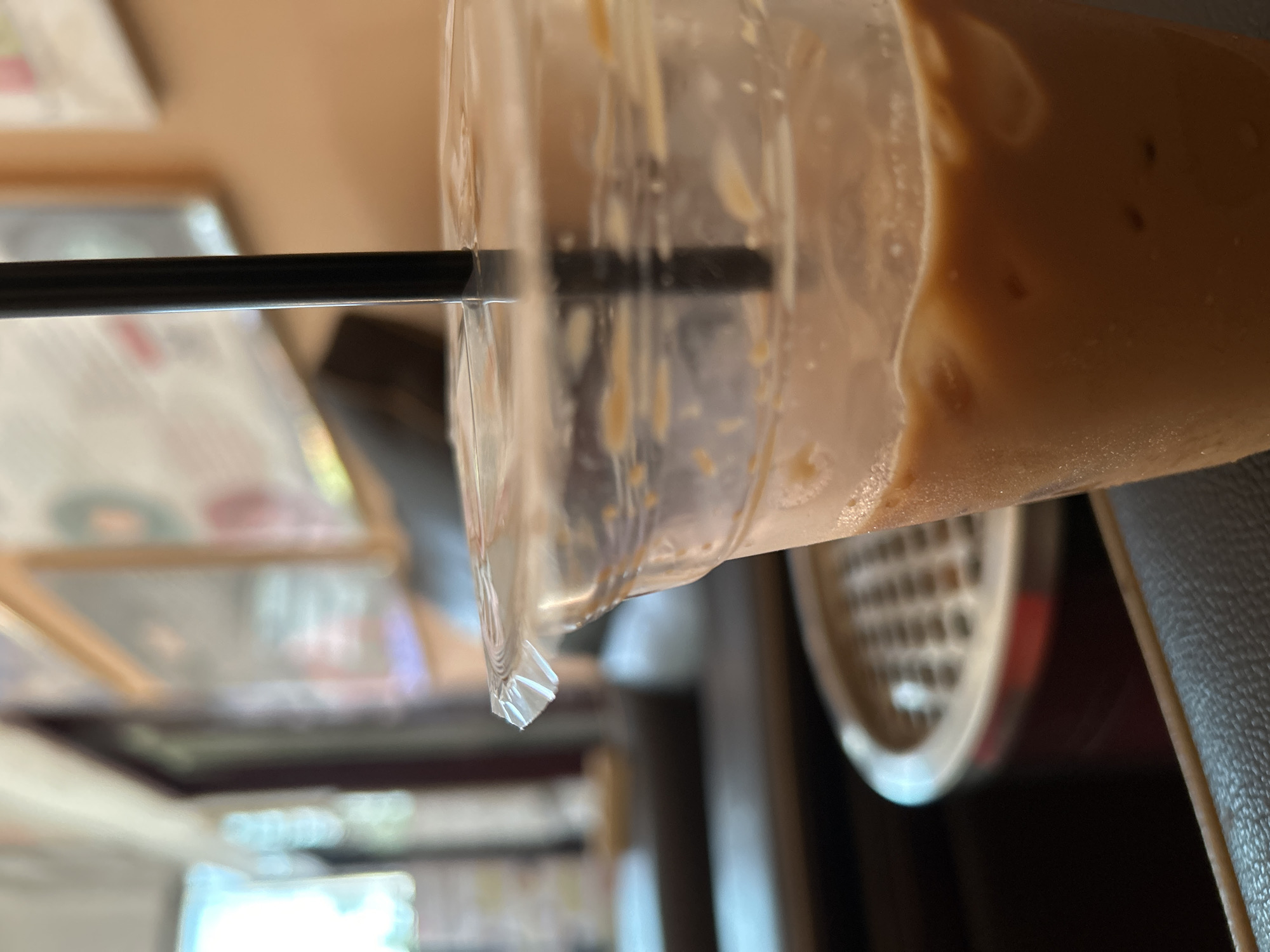 An upright close-up shot features a clear plastic cup filled with a light brown, creamy beverage, likely an iced coffee or milky tea. A black plastic straw is inserted into the drink, which appears partially consumed, with some liquid residue clinging to the inside of the cup. Small, dark speckles are visible within the drink itself.
The background is intentionally blurred, indicating a shallow depth of field. To the left, out-of-focus shapes and colors suggest the presence of printed menus or posters, featuring hues of green, pink, and brown. To the right, a dark, possibly upholstered surface, such as a chair or booth, is visible, along with a blurry, circular object exhibiting a grid-like pattern, possibly a fan or speaker.
The overall setting appears to be indoors, likely a cafe or restaurant in Lhokseumawe, Indonesia, during the daytime, given the soft, ambient lighting. No specific activity is depicted other than the presence of the drink. There is no visible text in the image.