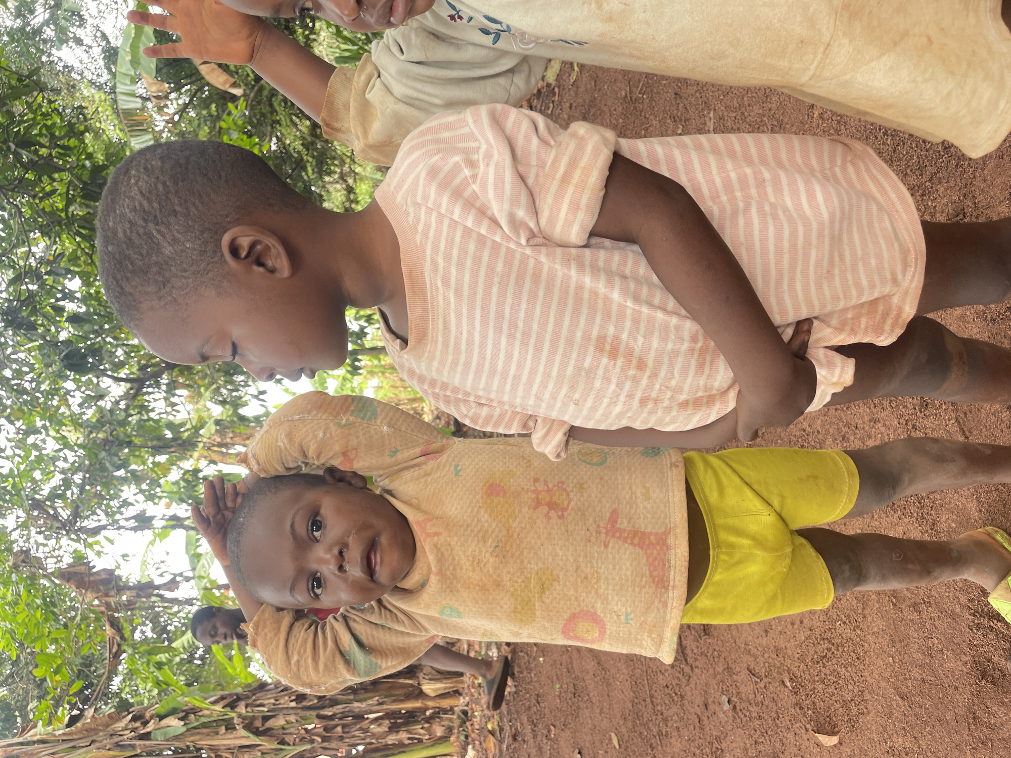 The image depicts a group of children in what appears to be a rural area of Ogbe, Nigeria. The ground is reddish-brown, possibly dirt or clay. There are some trees and vegetation in the background. Three children are prominently visible: One child is wearing a light-colored shirt with animal prints and bright yellow shorts. The second child is wearing a pink and white striped shirt. The third child is wearing a neutral colored dress.
