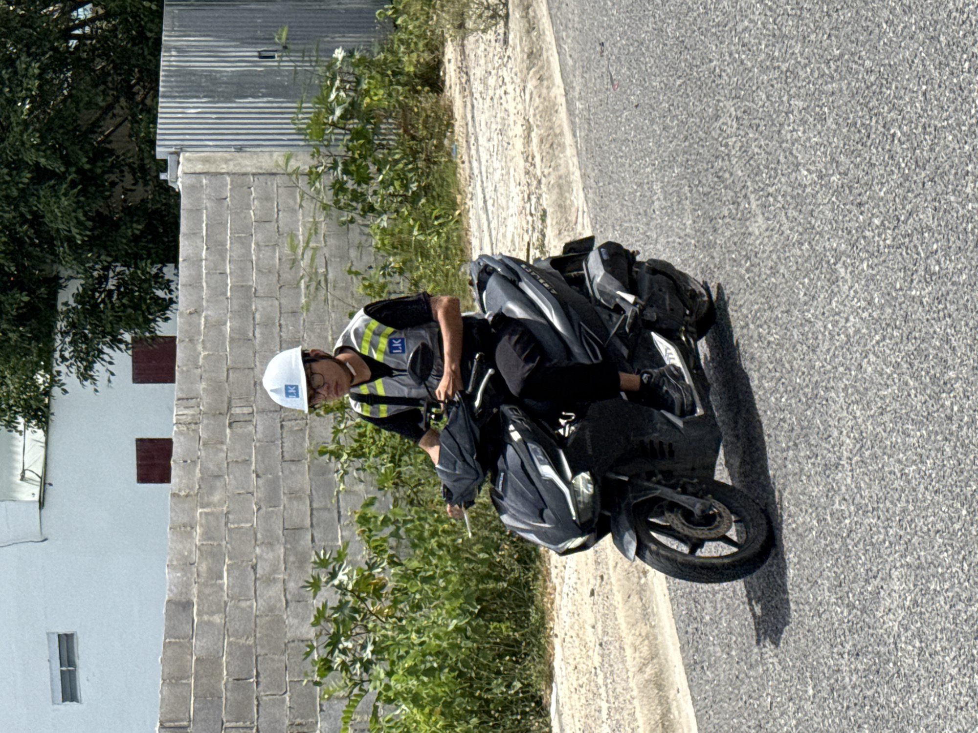 A person wearing a white hard hat and a reflective vest is riding a black scooter on a paved road. The person has short dark hair and is wearing glasses. They are dressed in a black shirt and black pants, and dark colored sneakers. The scooter has a black body with some silver accents. In the background, there is a low brick wall and a white building with two dark rectangular windows. Further in the background, there are green bushes and trees, and a corrugated metal fence. The scene appears to be in an urban or suburban area.