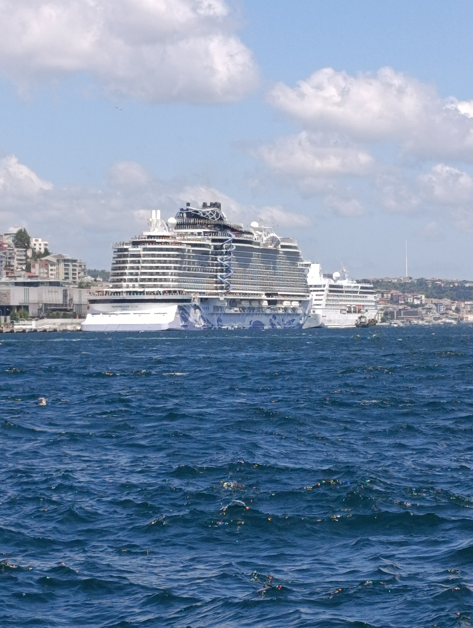 A large white cruise ship is docked in a harbor in Istanbul, Turkey. The ship has many decks with rows of windows and a distinctive, modern superstructure. The ship's hull is adorned with a large, colorful blue and white graphic design. In the background, buildings of various sizes and architectural styles line the shore, interspersed with trees. The water in the foreground is a deep blue, with visible waves and some scattered debris or litter floating on the surface. The sky above is mostly blue with scattered white clouds.
