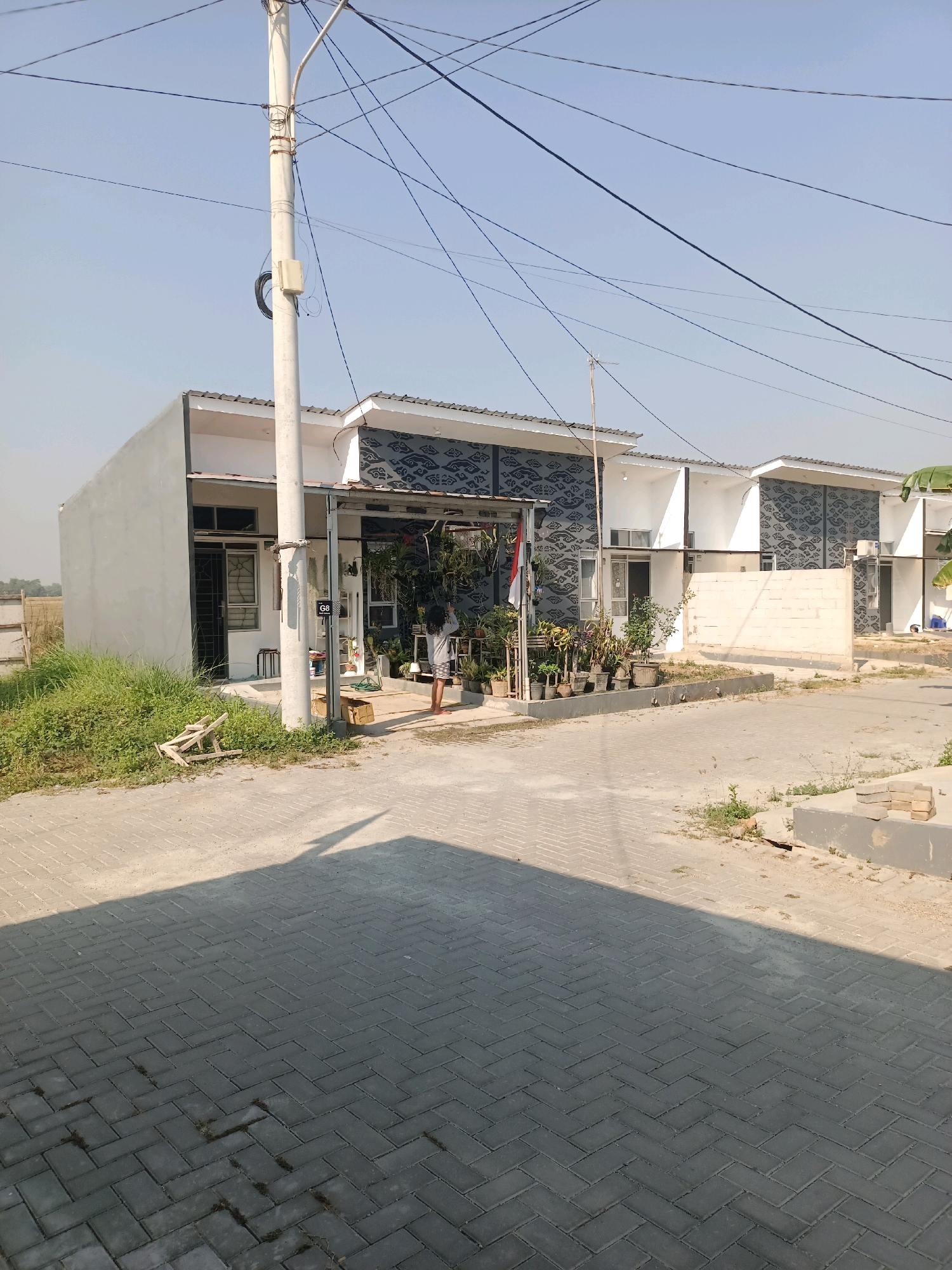 This image depicts a street scene in Karawang, Indonesia, featuring modern residential houses under a clear blue sky. The houses have white walls and dark patterned accents above the windows and doorways, with gray tiled roofs. A utility pole stands prominently in the foreground, adorned with numerous wires. In front of one of the houses, a person is standing near a collection of potted plants. The street is paved with interlocking gray bricks, and a shadow from a nearby structure stretches across it. To the left, there's a patch of tall green grass and some discarded wooden debris. The overall environment suggests a newly developed residential area.