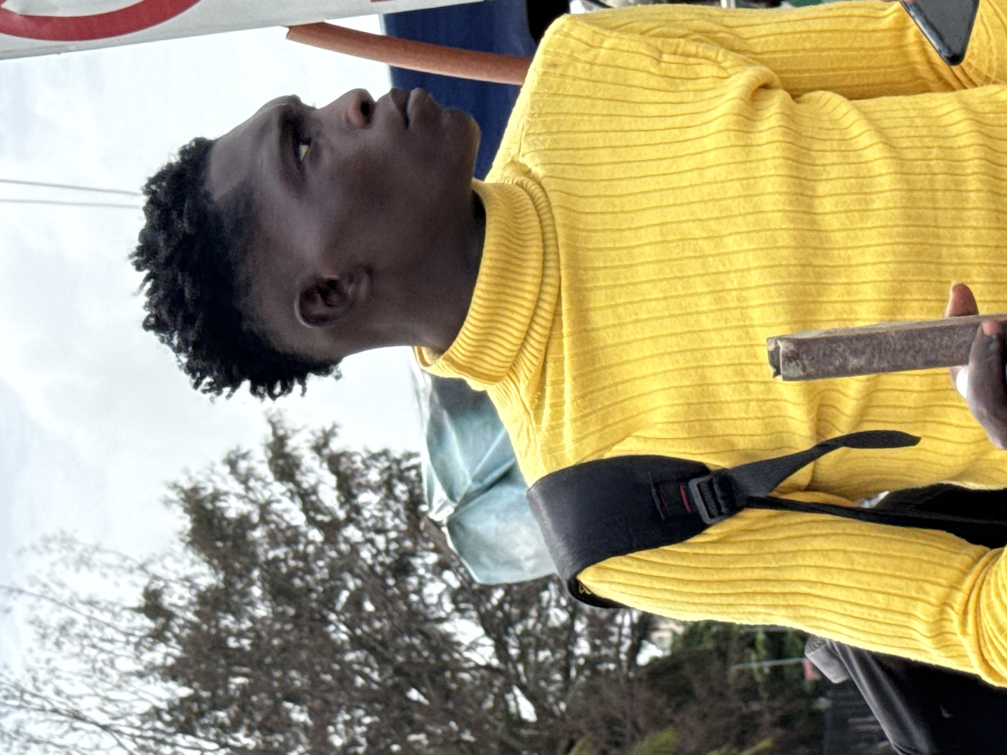 A young Black man, possibly in his late teens or early twenties, stands outside in what appears to be a public area, potentially a marketplace. He is looking upwards and to his left. He wears a yellow turtleneck sweater. A black strap is visible diagonally across his upper chest. The man is holding a dark, rectangular object in his right hand. The background contains bare tree branches, suggesting it may be winter or early spring. Elements of signage are partially visible at the upper left of the frame. Given the request to place this in Itirin, Nigeria, the scene suggests a daily life scenario of a young man in an urban setting.