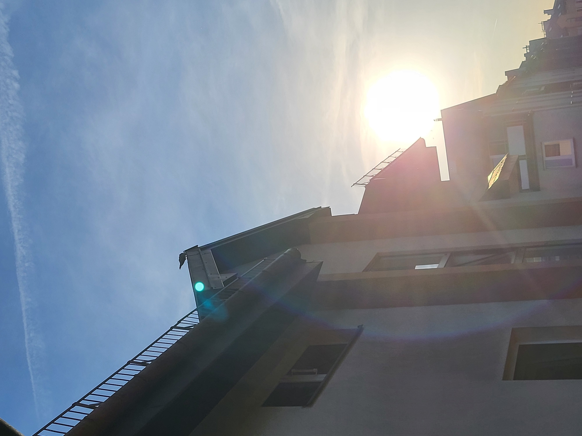 A low-angle shot looks up at a multi-story building against a blue sky with a bright sun. A long, thin cloud trail is visible in the upper left corner of the sky. The building appears to be modern, with windows and a flat roof. The sun is positioned behind the building, causing a glare. The location is Unknown, Unknown.