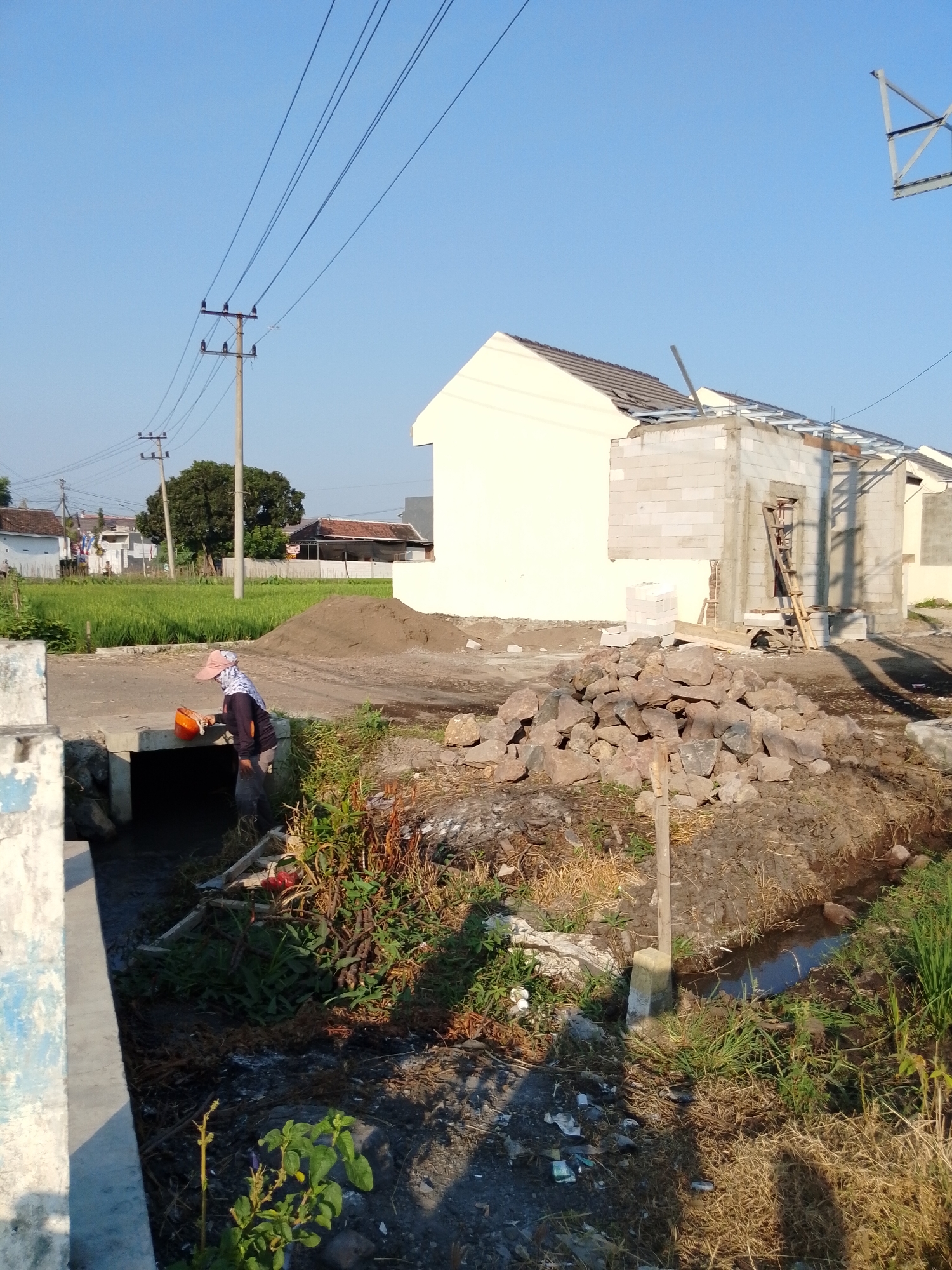 In Pasuruan, Indonesia, a construction site is depicted under a clear blue sky. A partially constructed house, made of cinder blocks and with a framed roof structure, stands to the right of the image. A pile of rocks sits in the foreground, to the left of a small, muddy ditch where a person, wearing a pink cap and dark clothing, is reaching into the ditch with an orange bucket. Green rice fields are visible in the background, with some houses and utility poles dotting the landscape. The ground is a mix of dirt, mud, and dry grass, with some scattered debris.