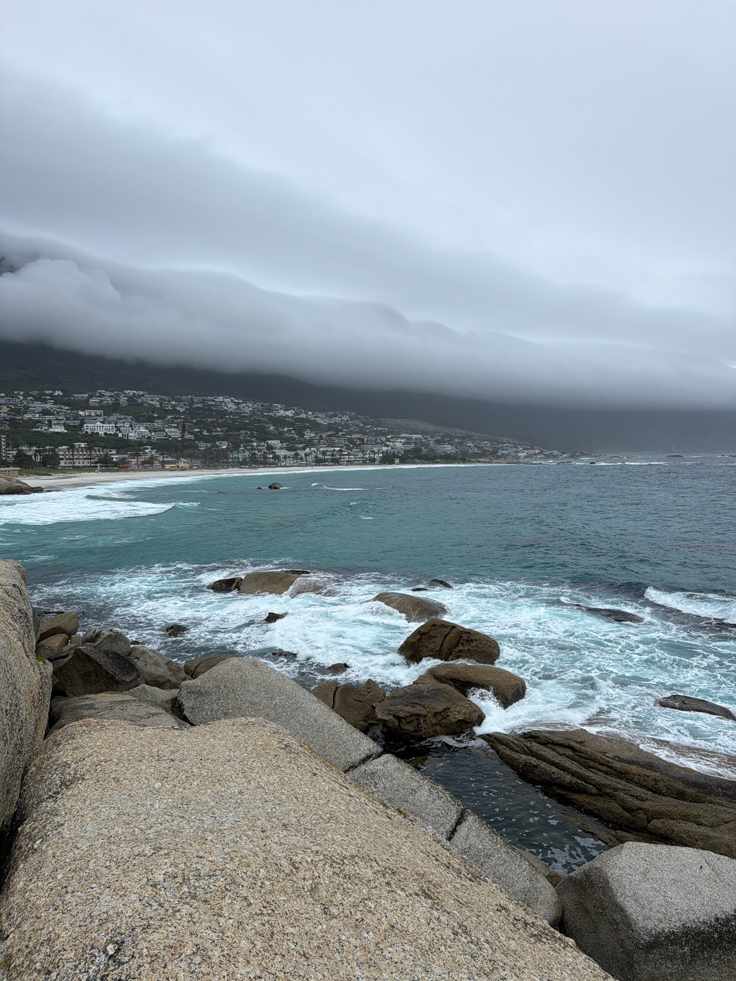 The image shows a cloudy, overcast view of the coastline at Camps Bay in Cape Town, South Africa. In the foreground, large granite boulders are visible, with smaller rocks and waves crashing against them. The middle ground features a sandy beach with a line of white buildings and palm trees, characteristic of the Camps Bay beachfront. Beyond the beach, the turquoise ocean stretches out, with white-capped waves rolling towards the shore. In the background, the Twelve Apostles mountain range is shrouded in thick, low-hanging clouds, obscuring the upper portions of the mountains and the sky. The overall impression is one of a dramatic, somewhat moody coastal scene due to the weather conditions. There are no people or animals visible in the image.
