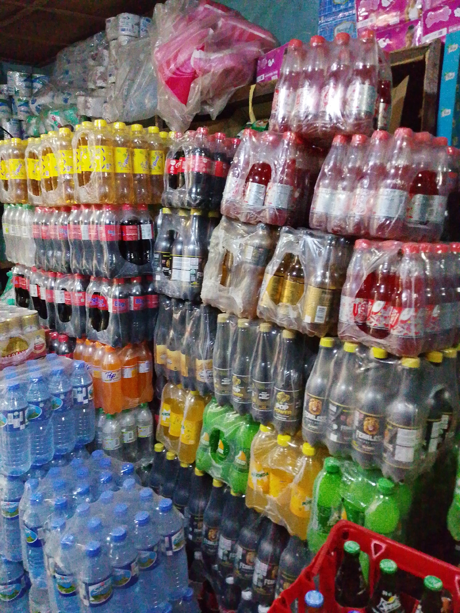 The image depicts a cluttered store shelf in an unknown location. Stacks of bottled drinks, mostly soft drinks and water, dominate the scene. They are arranged in multi-packs, often wrapped in clear plastic. Various brands and flavors are visible, including Coca-Cola, orange soda, lime soda, and others. In the background, more packaged goods are stacked. The store appears to be small, with limited space.
