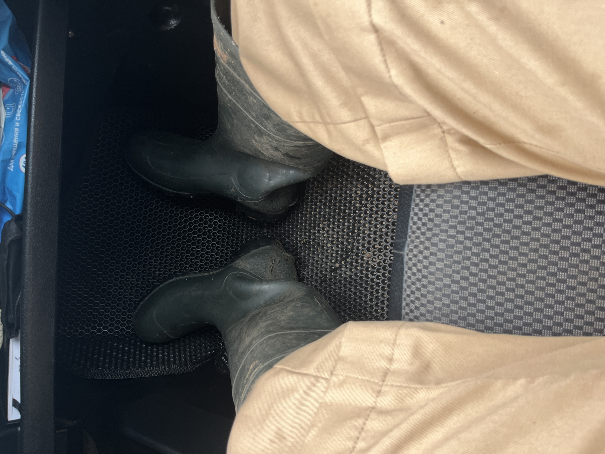 The image shows a pair of dark green rubber boots on a person's feet, standing on a textured dark gray floor mat inside what appears to be a vehicle. The person is wearing light beige pants. The boots are slightly dirty. In the background, there is a dark surface, possibly the dashboard or console of the vehicle, with a blue and white package resting on it. The floor mat has a hexagonal pattern in some areas and a checkered pattern in others. The overall scene is indoors, likely within a car, based on the visible elements of the interior. The location of Kemul, Russia, is not discernible from the visual information in the image.