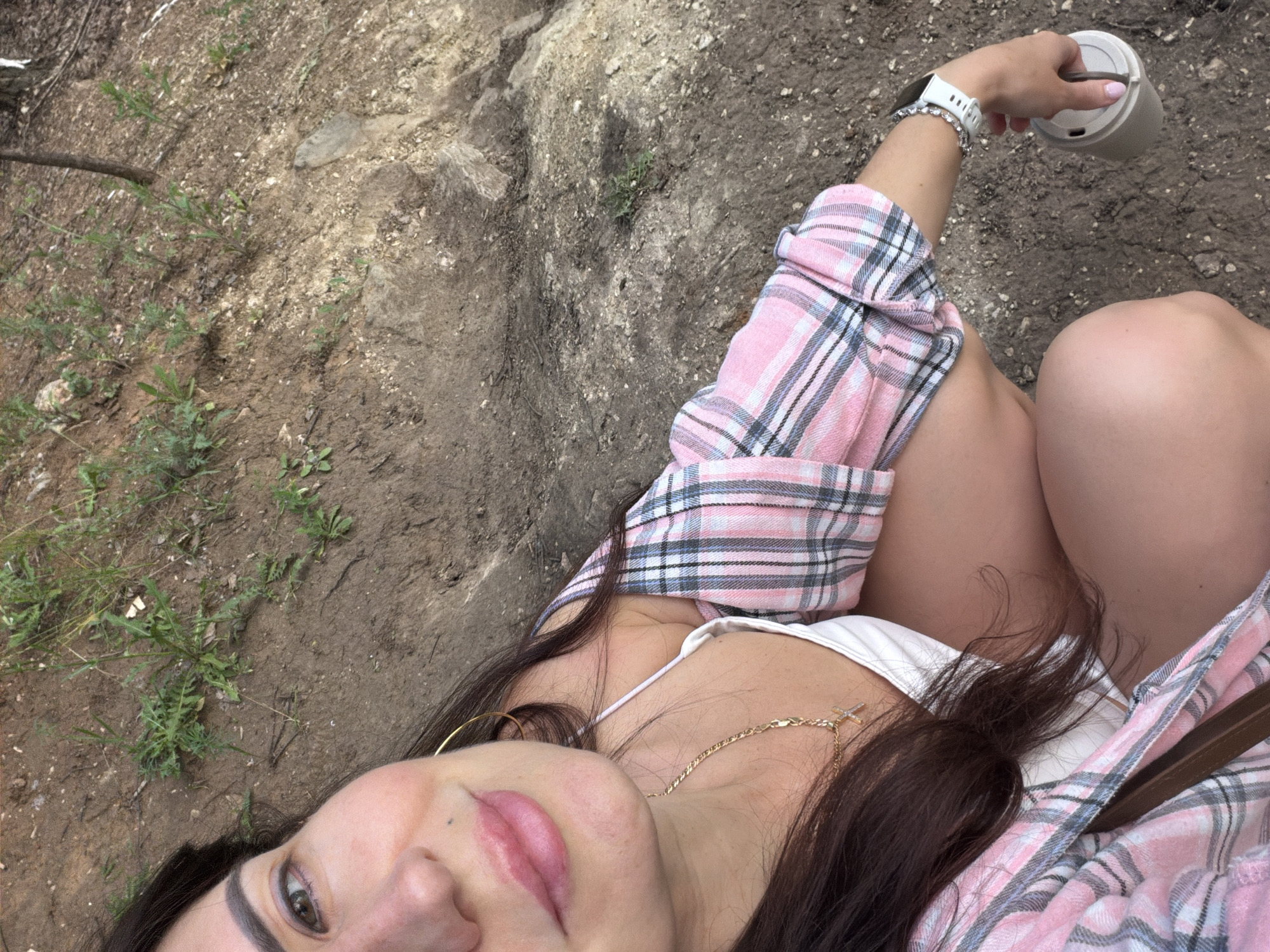 A woman with dark hair and light skin is crouching outdoors, holding a grey travel mug in her right hand. She is wearing a white top under a pink and blue plaid shirt and a silver bracelet on her left wrist. The background is a dirt slope with sparse green vegetation and rocks. The location is the city of Volzhskiy, Russia.