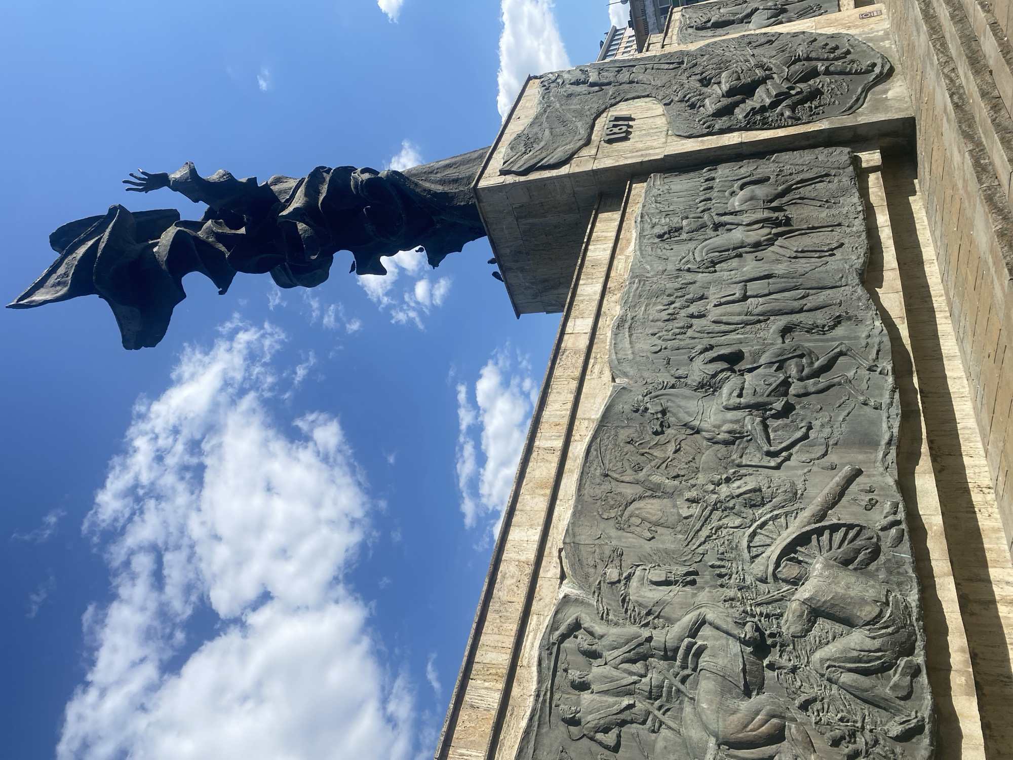 The image shows the "Mihai Eminescu" monument in Iași, Romania, under a bright blue sky with scattered white clouds. The monument features a towering, abstract bronze sculpture of a figure with one arm raised, and a large relief panel depicting historical scenes. The relief panel is made of dark metal and shows a procession of figures on horseback and on foot, as well as a cannon. The monument is situated on a stone base with steps leading up to it. The overall scene is one of historical commemoration and public art in an urban setting.
