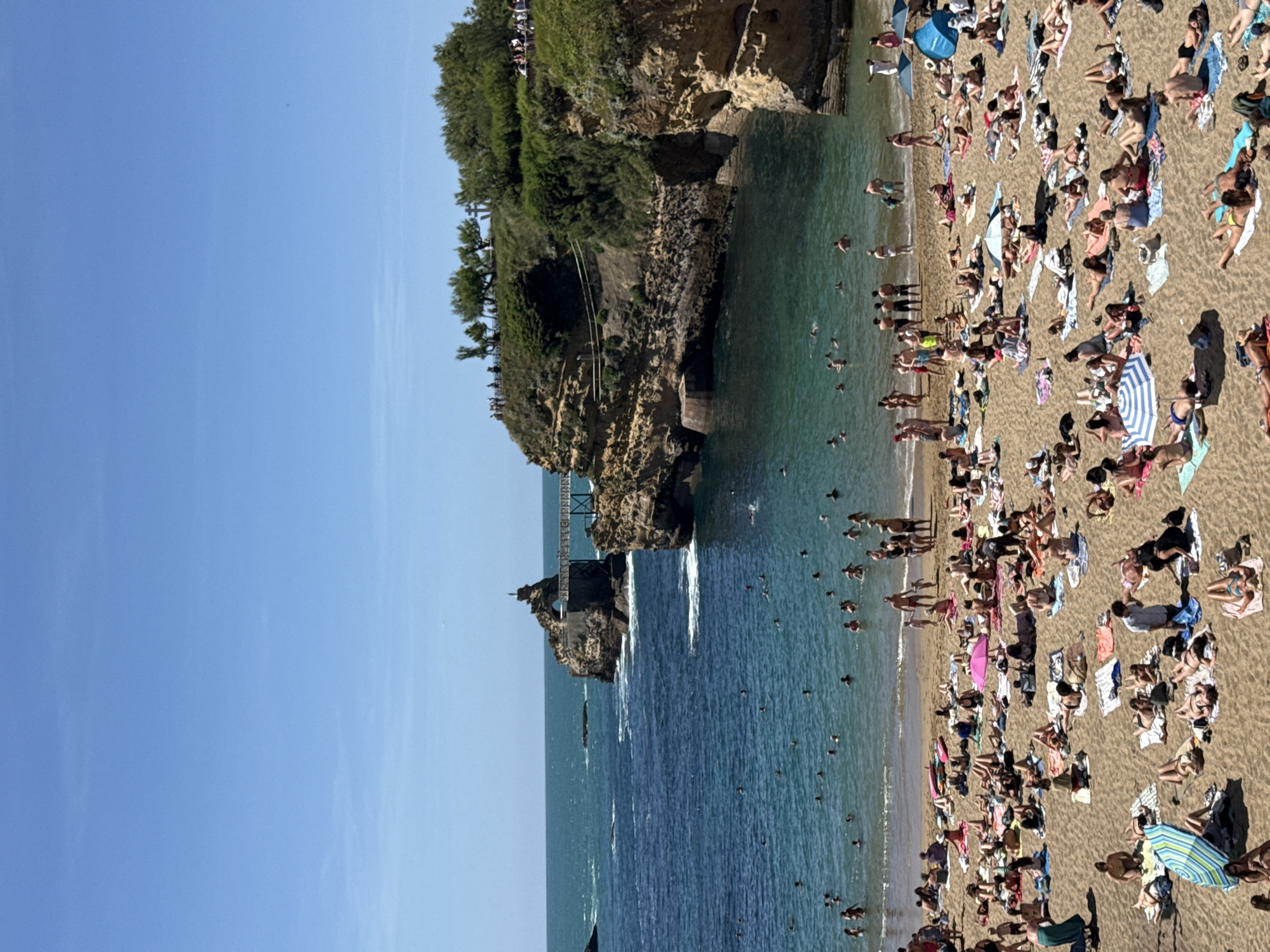 The image depicts a crowded beach scene on a sunny day, likely in Biarritz, France, given the distinctive rock formations. The foreground is dominated by a sandy beach filled with numerous people sunbathing, lying on towels, and relaxing under umbrellas. Many individuals are in swimwear, with some visible in bikinis and swim trunks. A few people are standing or walking on the sand.
In the middle ground, the turquoise ocean is populated by many swimmers, some wading in the shallow water while others are further out. Waves are gently breaking near the shore.
The background features dramatic, rocky cliffs that jut out into the sea, characteristic of the Biarritz coastline. A distinctive rock formation with a bridge connecting it to the mainland is prominently visible. Several trees and vegetation grow on top of the cliffs, and a group of people can be seen observing the scenery from a vantage point on the cliff edge. The sky is a clear, bright blue, indicating fair weather. The overall impression is a vibrant and lively beach environment, popular with tourists and locals alike.