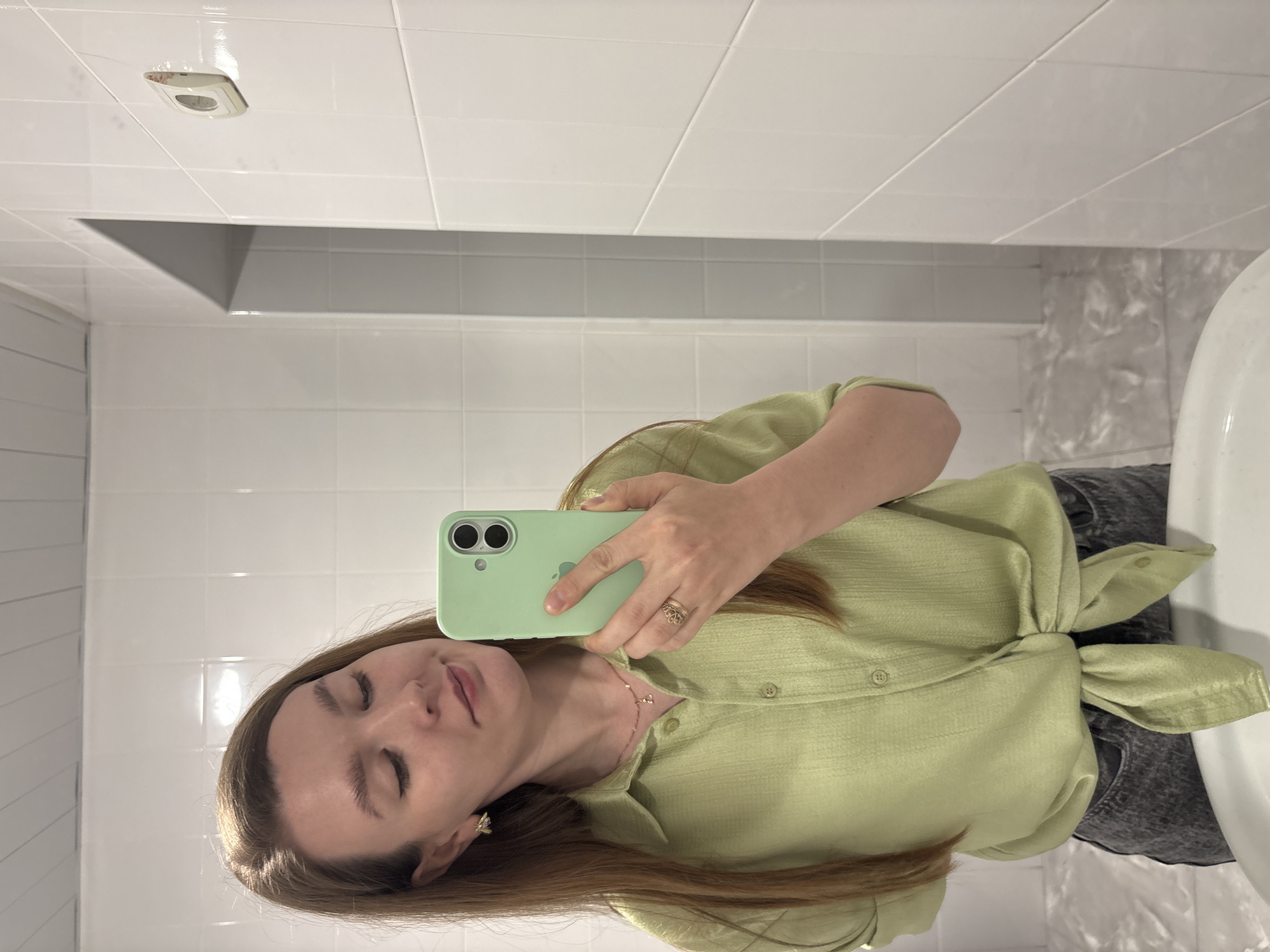A woman is taking a selfie in a bathroom. The bathroom is tiled in white and gray. There is a vent in the ceiling. The woman is wearing a light green shirt with a knot at the waist and gray jeans. She is holding a light green phone in front of her face. The location is a bathroom, presumed to be in Izhevsk, Russia.