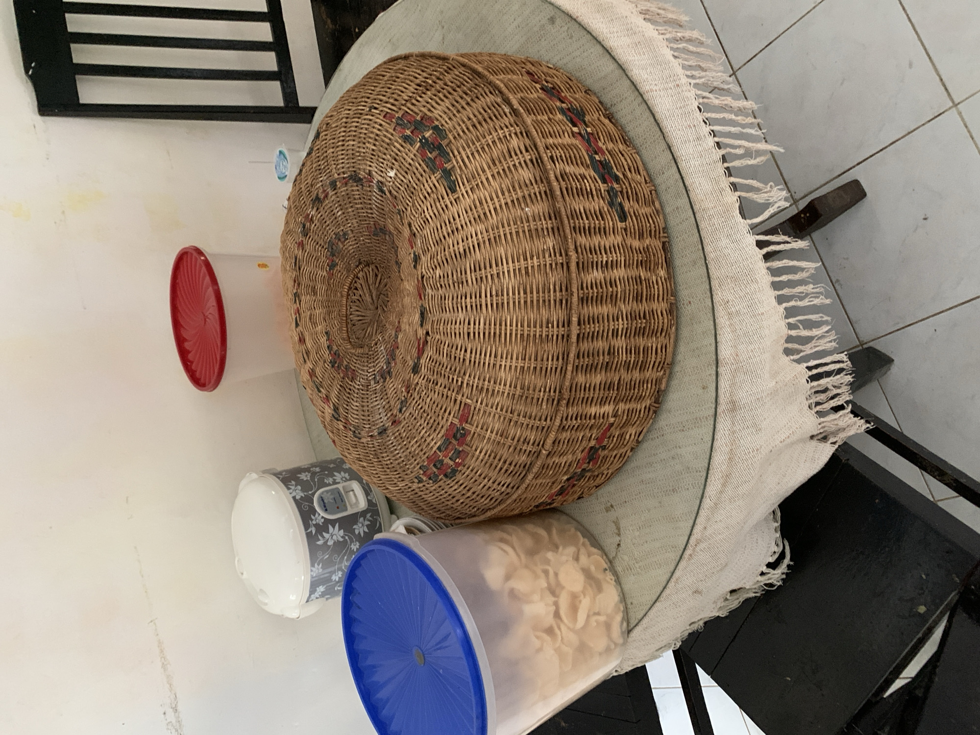 The image depicts a round, woven basket with decorative red and blue patterns, placed on a glass-topped table covered with a light-colored, fringed cloth. To the left of the basket is a clear plastic container with a blue lid, filled with light-colored chips. Behind the container is a white rice cooker with a floral pattern. To the right of the basket, a white plastic container with a red lid is visible. A black chair is partially visible on the right side of the frame, and on the far left, another black chair leg is present. The scene appears to be indoors, with a light-colored wall and tiled floor. The overall impression is that of a domestic setting in a home or food establishment. The items on the table suggest food storage and preparation. The image does not provide specific details that would definitively place it in Uleepulo, Indonesia, beyond the provided context.