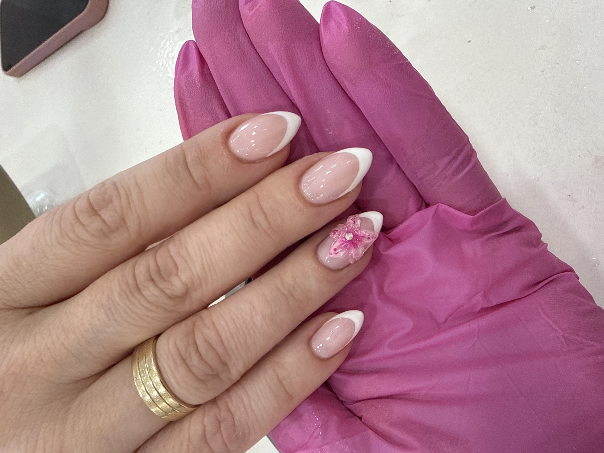 A close-up shot shows a hand with manicured nails, featuring a classic French manicure with white tips and a natural pink base. The ring finger has a pink floral embellishment. The hand is adorned with a gold ring on the ring finger, and it rests on a pink gloved hand. The background consists of a speckled white surface, possibly a table or countertop, with a blurred object in the upper left corner. This image could potentially be taken in a nail salon or similar setting in Sofia, Bulgaria.