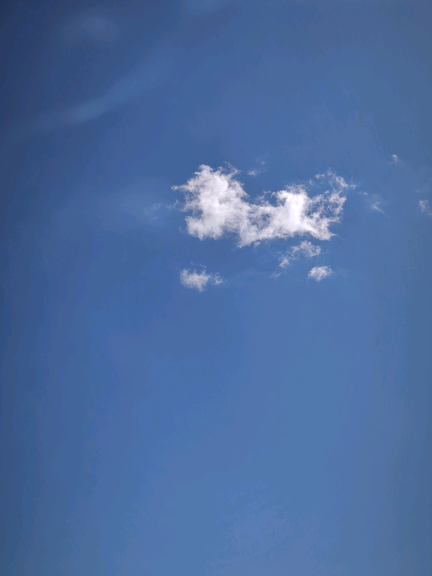 A photograph captures a bright blue sky with a few white clouds. One cluster of clouds is centrally located, appearing fluffy and somewhat dispersed. Additional smaller clouds are visible in the lower portion of the image. The scene is set in the city of Karachayevsk, Russia, a location known for its mountainous terrain and clear skies, especially during favorable weather conditions.