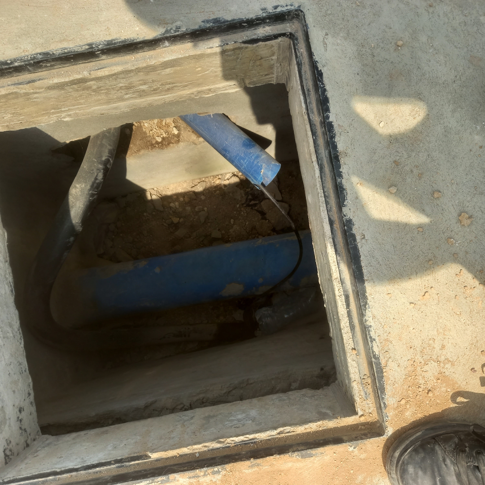 In Bububu, Tanzania, a rectangular concrete structure with a black-lined opening is visible, revealing an underground space. Inside this space, two large blue pipes are present, one positioned horizontally and another angled downwards. A thick black cable runs from the angled pipe towards the viewer, disappearing into the darkness. The surrounding area is a mix of concrete and dirt, with some debris scattered around. The light casts a distinct shadow on the concrete surface, suggesting it is daytime. A black shoe is visible in the lower right corner of the image, indicating the presence of a person or persons at the scene, likely observing or working in the underground area.