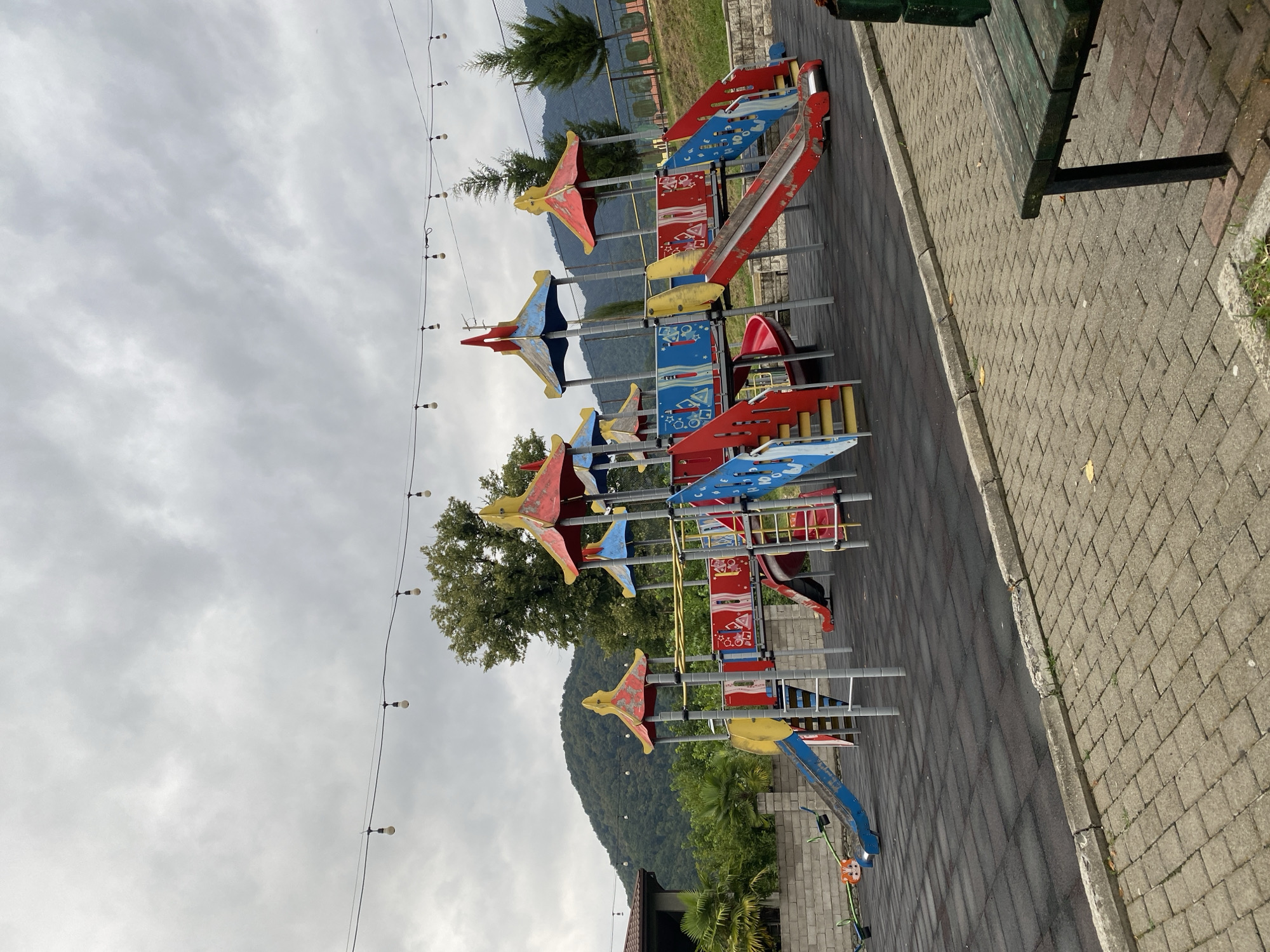 The image depicts a playground in Lagodekhi, Georgia, under a cloudy sky. The playground features a multi-colored structure with slides, stairs, and platforms. A seesaw is visible on the left side of the playground. The ground is covered with black rubber mats, and the surrounding area has a paved walkway with brick pavers and a concrete curb. In the background, there are trees and rolling hills, indicative of a natural landscape. String lights are suspended overhead. A wooden bench is positioned on the right side of the paved area.