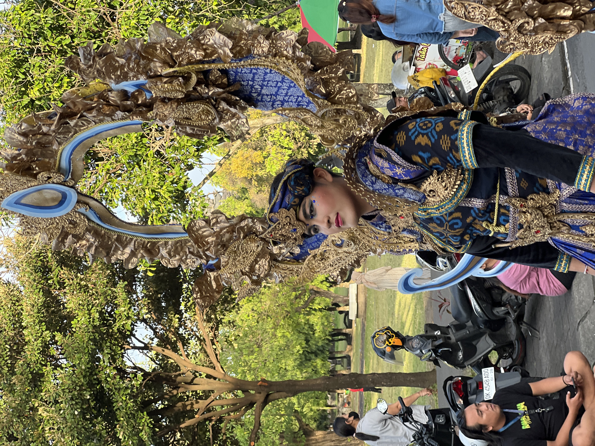 In Denpasar, Indonesia, a person adorned in a vibrant, elaborate costume stands in what appears to be a public park. The costume features gold, blue, and black fabric with intricate patterns, including a large, ornate headdress and shoulder adornments that resemble wings. The individual has dramatic makeup, with blue eyeshadow and dark eyeliner. They are looking to the left of the frame.
In the background, several motorcycles are parked. One is a black scooter with a yellow and black helmet. Another is a white scooter. A man in a black t-shirt is sitting cross-legged on the ground in the foreground, looking towards the seated person. To the right, a woman in a denim jacket stands. In the middle ground, other people are visible, including a person in a light-colored hijab. The environment is outdoors, with lush green trees and a grassy area, suggesting a park or an open public space. The lighting indicates it is daytime, likely late afternoon given the warm light.