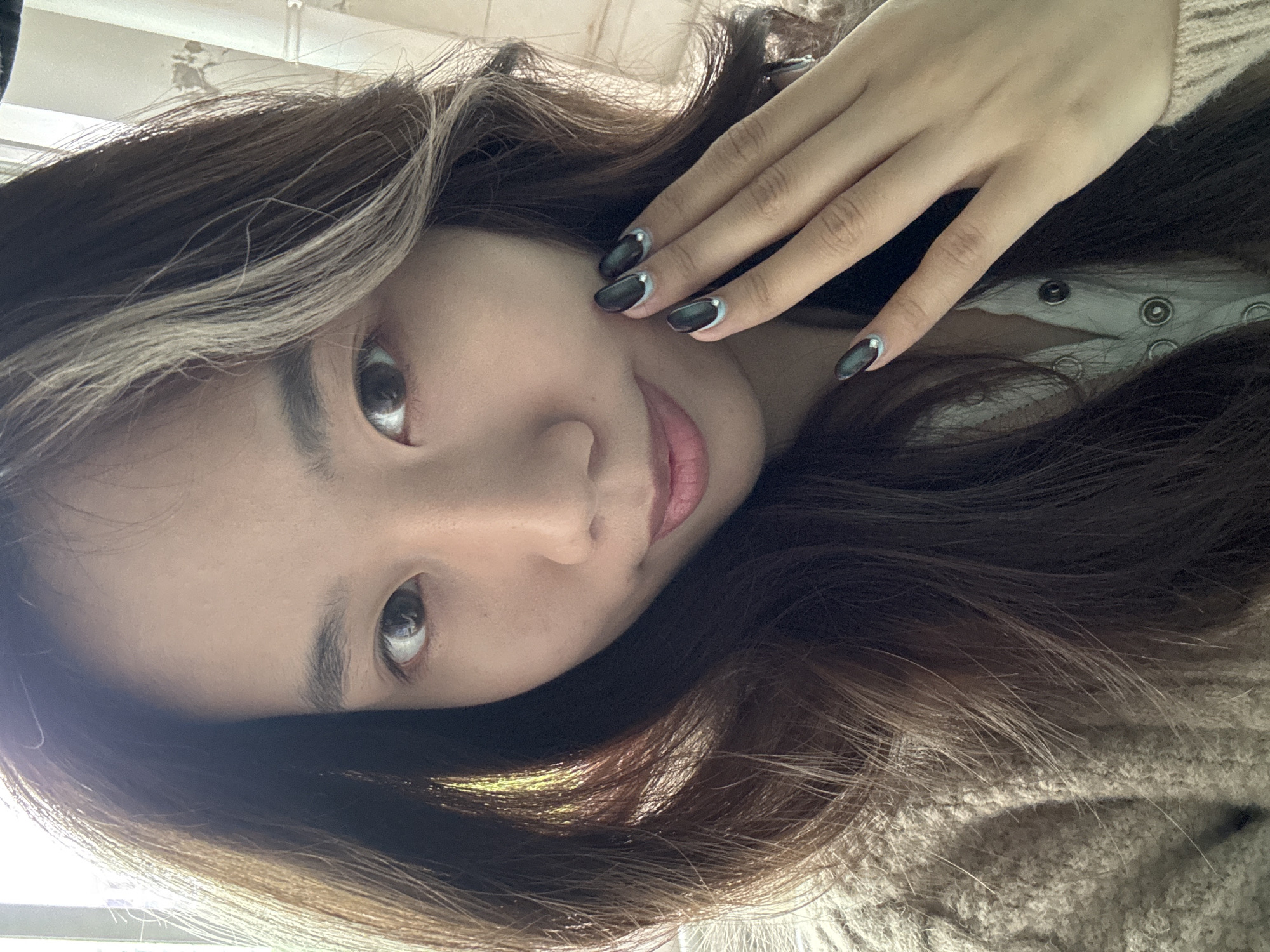 The image presents a close-up, frontal portrait of a single East Asian female subject, captured indoors in Nan'an District, China. The subject, appearing to be in her 20s or 30s, looks directly into the camera with dark brown eyes and a slight, closed-lip smile. Her long, dark brown hair, featuring prominent lighter, possibly blonde or greyish, streaks on the left side, frames her face and flows down her shoulders.
The subject's left hand is positioned near the lower left side of her face, fingers gently curved. Her fingernails are manicured with a dark, glossy, possibly black or very dark grey polish. Each nail displays a light blue or silver crescent-shaped line at its base, accompanied by a small, spherical silver embellishment centrally located within the crescent.
She is dressed in a light-colored, possibly cream or beige, knitted cardigan with a visible textured pattern. Beneath the cardigan, a light-toned undergarment with multiple silver-toned snap buttons is visible. The scene is illuminated by a strong, diffused light source originating from the upper left, casting highlights on her hair and face. The background is out of focus, revealing light-colored interior elements, possibly a wall or window with subtle indications of wear.