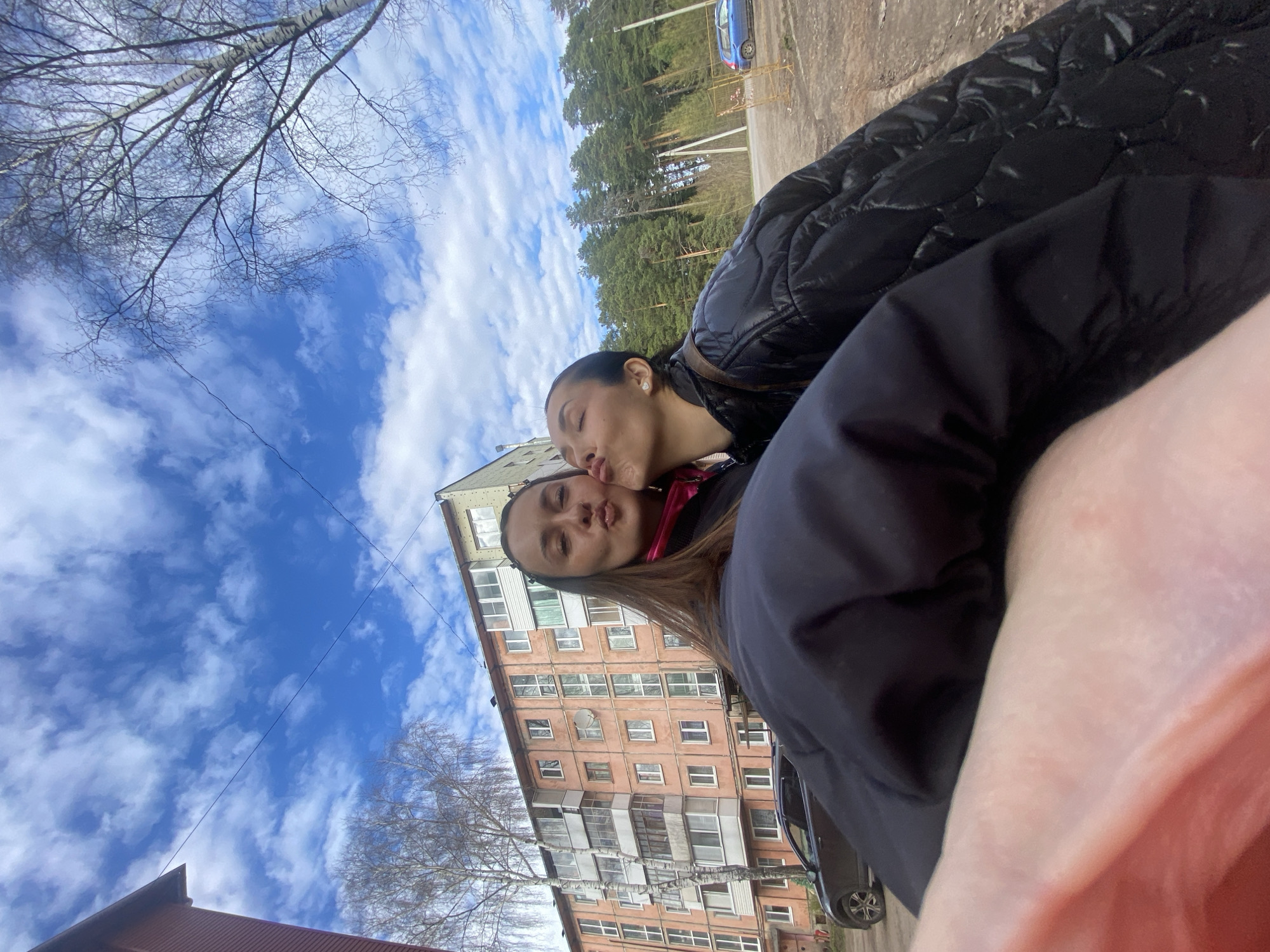 This outdoor selfie, taken in Votkinsk, Russia, captures a tender and playful moment between two young women on a bright, cloudy day. The image is significantly tilted clockwise.
In the foreground, one woman with dark hair, pulled back, and wearing a black puffer jacket, is seen with her eyes closed, leaning in to kiss the cheek of the second woman. She has a small white earring visible. The second woman, with lighter brown hair and a pink or red collar visible beneath a dark jacket, looks towards the camera with slightly puckered lips, possibly in a playful response to the kiss. Part of the arm or hand of the person holding the camera is visible in the bottom right foreground.
The background features a multi-story residential building, likely an apartment block, with a reddish-brown facade, numerous windows, and balconies. Bare tree branches, possibly birch or similar deciduous trees, are prominent against the sky in the upper left and lower central parts of the image. A line of tall pine trees borders what appears to be a road or path on the right side, with a blue car partially visible parked there. The sky is a clear blue with scattered white clouds, indicating daytime. No visible text is present. The overall impression is one of affection and cheerfulness.