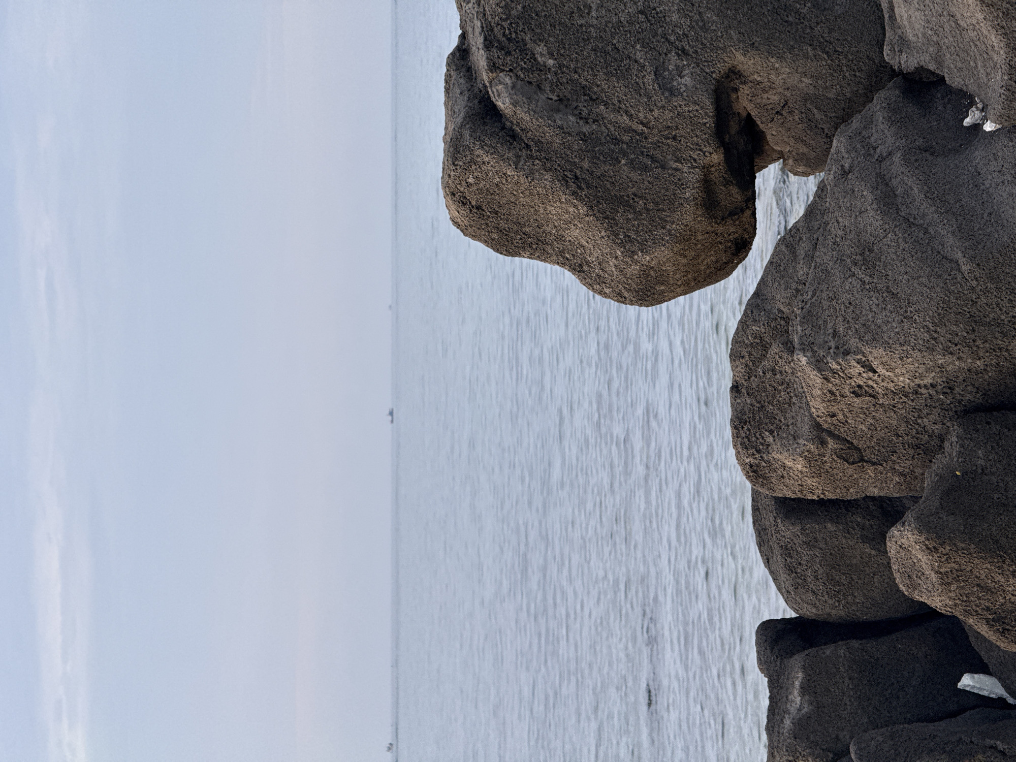 The image depicts a coastal scene with a calm ocean under a pale, overcast sky. Large, dark, porous rocks dominate the foreground, with a cluster of them on the left and a single, prominent rock formation on the right, which has a rounded, almost animalistic shape. The water is a muted blue-grey, with a gentle ripple pattern. In the distance, on the horizon, two small boats are visible. The overall lighting suggests it is either early morning or late afternoon. The location is identified as Lý Sơn Special Administrative Region, Vietnam, known for its volcanic rock formations.