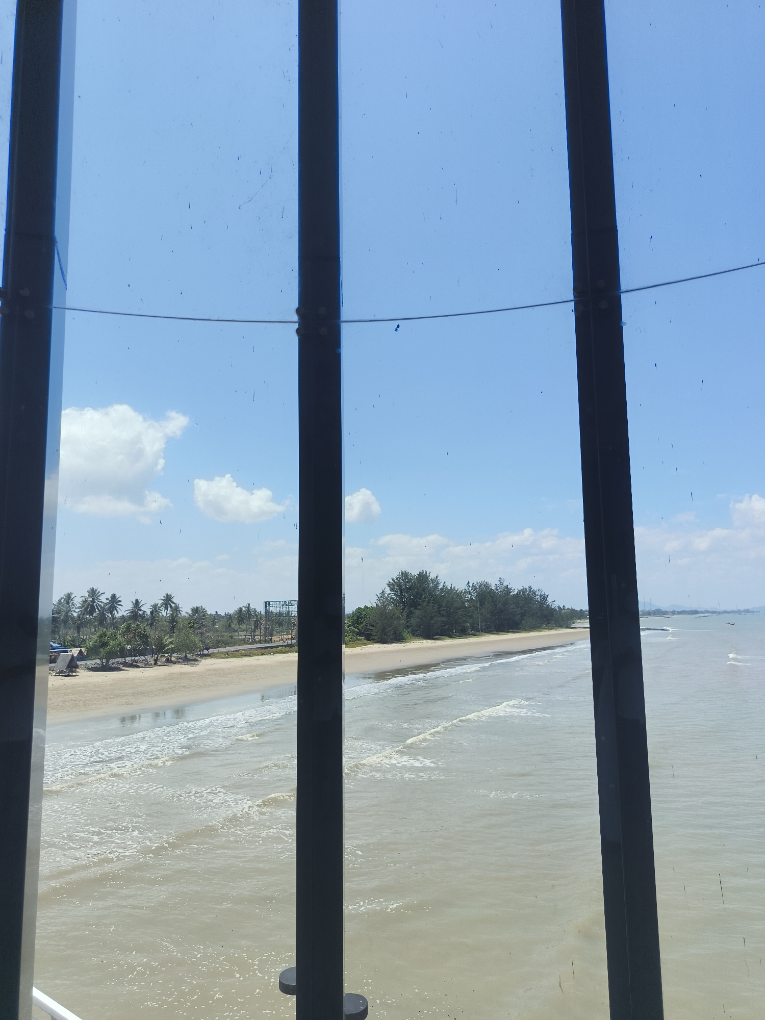 The image shows a view of a beach and the ocean through vertical bars. The beach is sandy with a line of trees and palm trees behind it. There are some small structures on the beach, including a hut. The ocean is calm with gentle waves. The sky is blue with some white clouds. The photograph appears to have been taken from a building or structure with a view of the coast. The location is identified as Beringin, Indonesia.