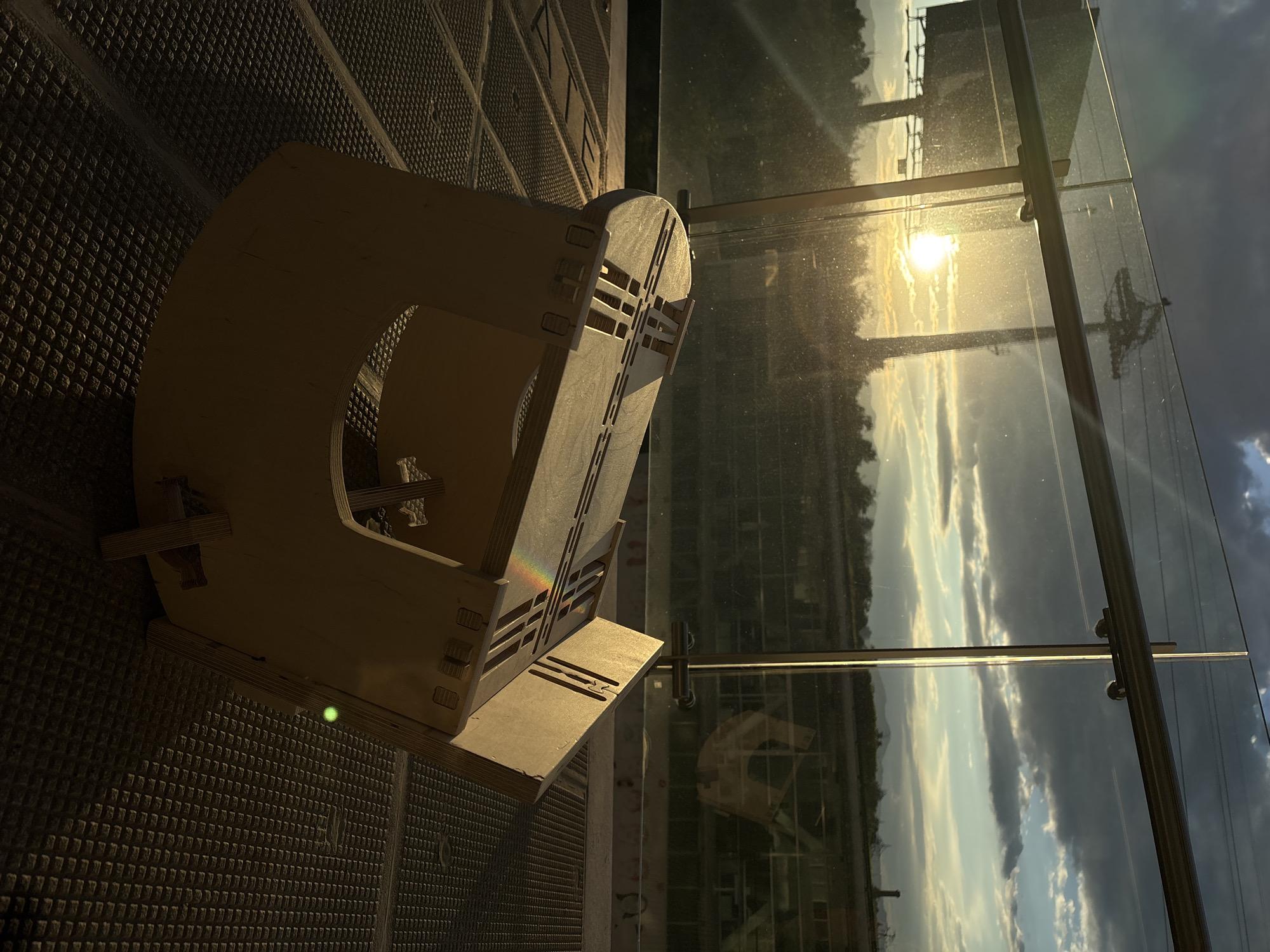 A wooden object is placed on a dark surface with a geometric pattern. Beyond it, there is a window reflecting the sky and a cityscape in the background. The sun is prominently shining, creating a bright spot in the sky. The location appears to be in Mexico City, Mexico. The reflection in the glass suggests a daytime scene with a mix of clouds and clear sky. The wooden object's design features a series of interconnected shapes and cutouts.