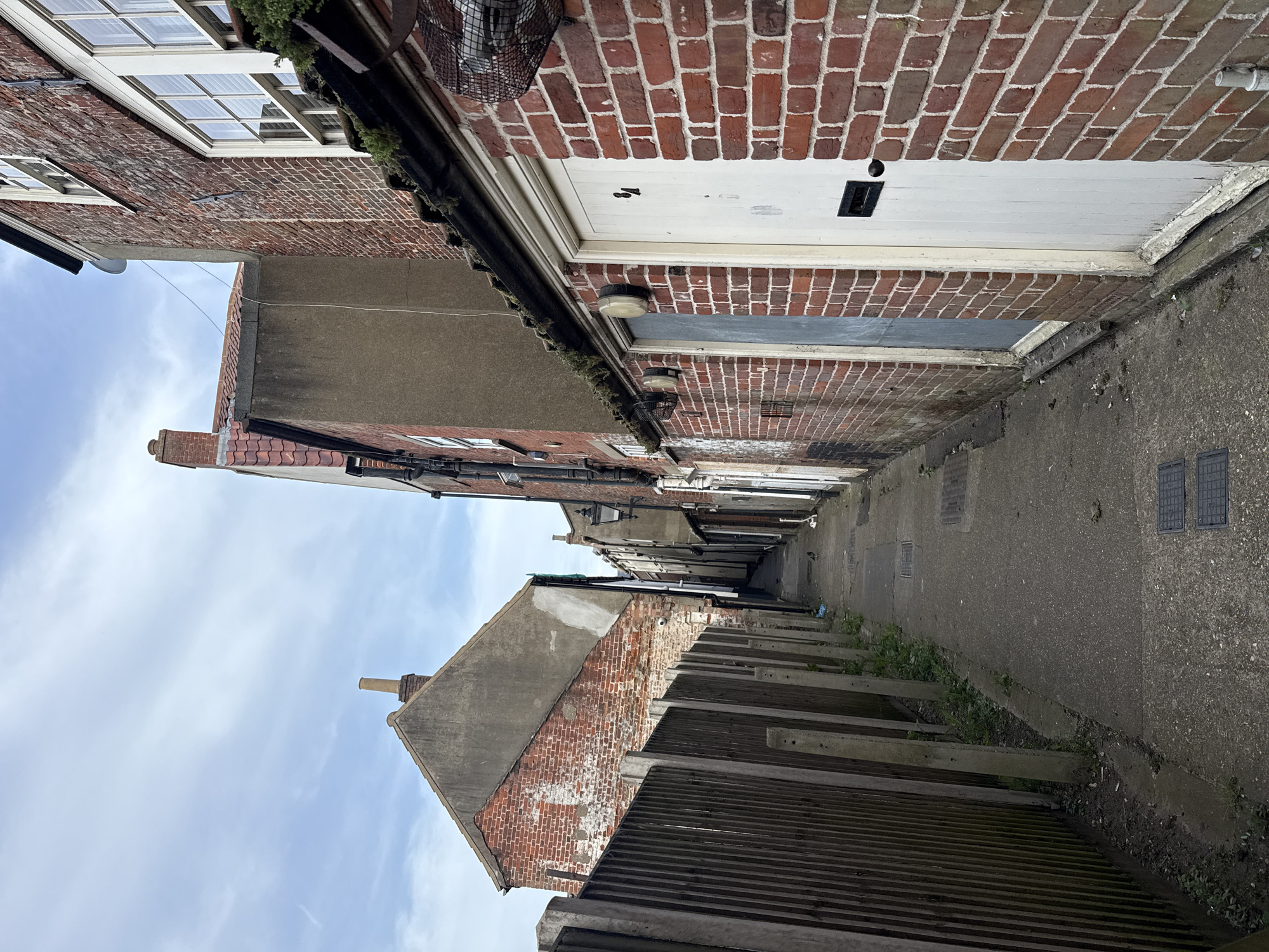 A narrow alleyway in Great Yarmouth, United Kingdom, is framed by brick buildings and a concrete walkway. The alley appears to recede into the distance, with buildings lining both sides. Some buildings have windows and doors, while others appear to be plain brick walls. The alleyway is concrete, with minor weed growth along the edges. A corrugated fence is on one side. The sky is visible at the top of the image.
