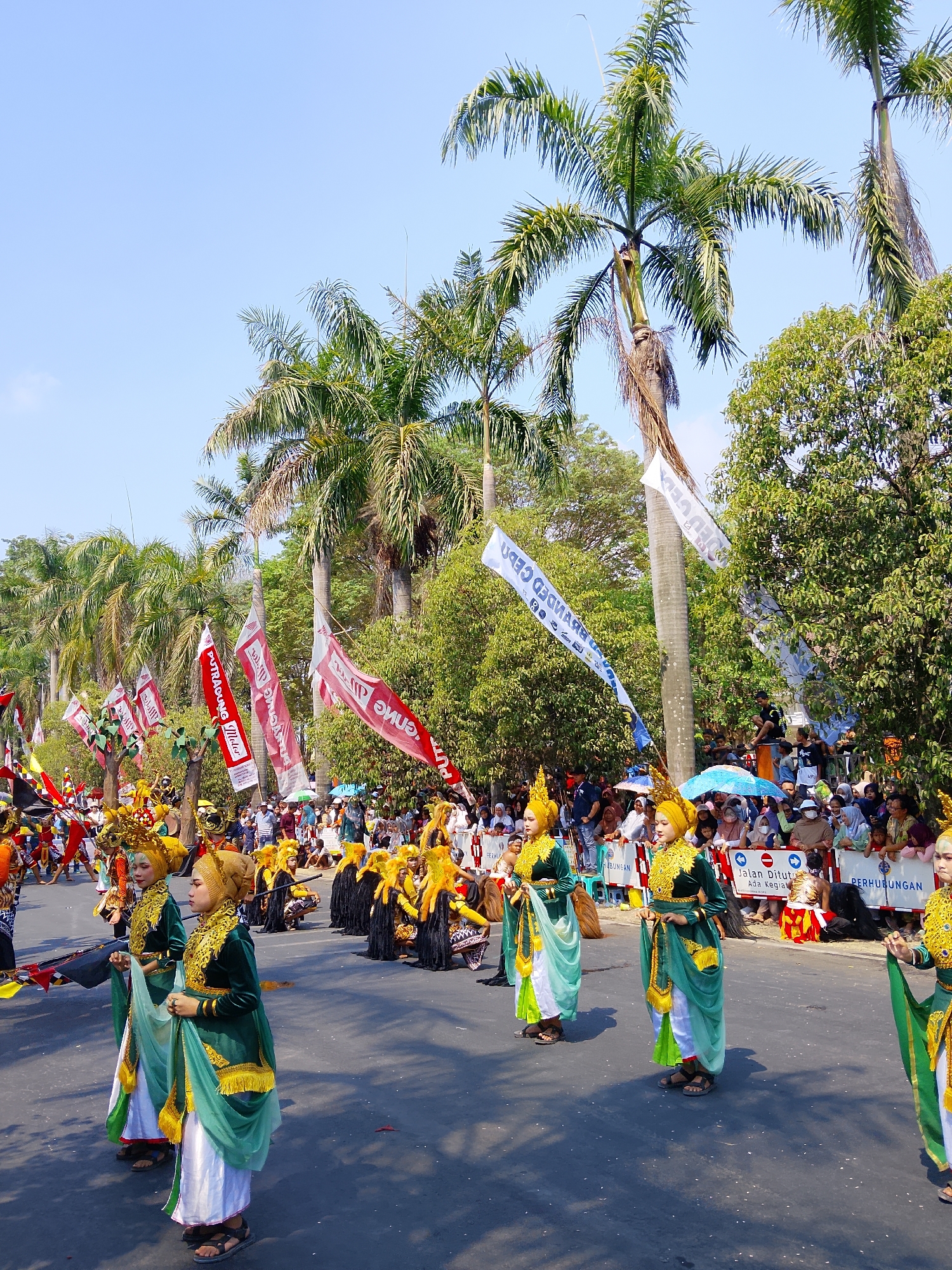 The image depicts a vibrant cultural parade in Balun, Indonesia, under a clear blue sky. The foreground features several performers in traditional attire, characterized by elaborate gold headdresses and ornate green and gold garments over white trousers. They are dressed in layered robes and flowing sashes, with one individual holding a green veil. Behind them, other performers are visible, some wearing black and gold costumes and headdresses, and others in orange and yellow.
The parade route is lined with spectators seated or standing behind barriers. Many onlookers are holding umbrellas, suggesting a sunny day. The background is dominated by tall palm trees and lush green foliage, indicating a tropical environment. Several flags are displayed along the parade route; some are red with white lettering, while others are white with blue and black lettering. Banners in the background read "PERHUBUNGAN" and "JALAN DITUTUP ADA KEGRA." The street itself is asphalt, and shadows are cast by the trees and structures, suggesting it is daytime. The overall scene conveys a festive and celebratory atmosphere.