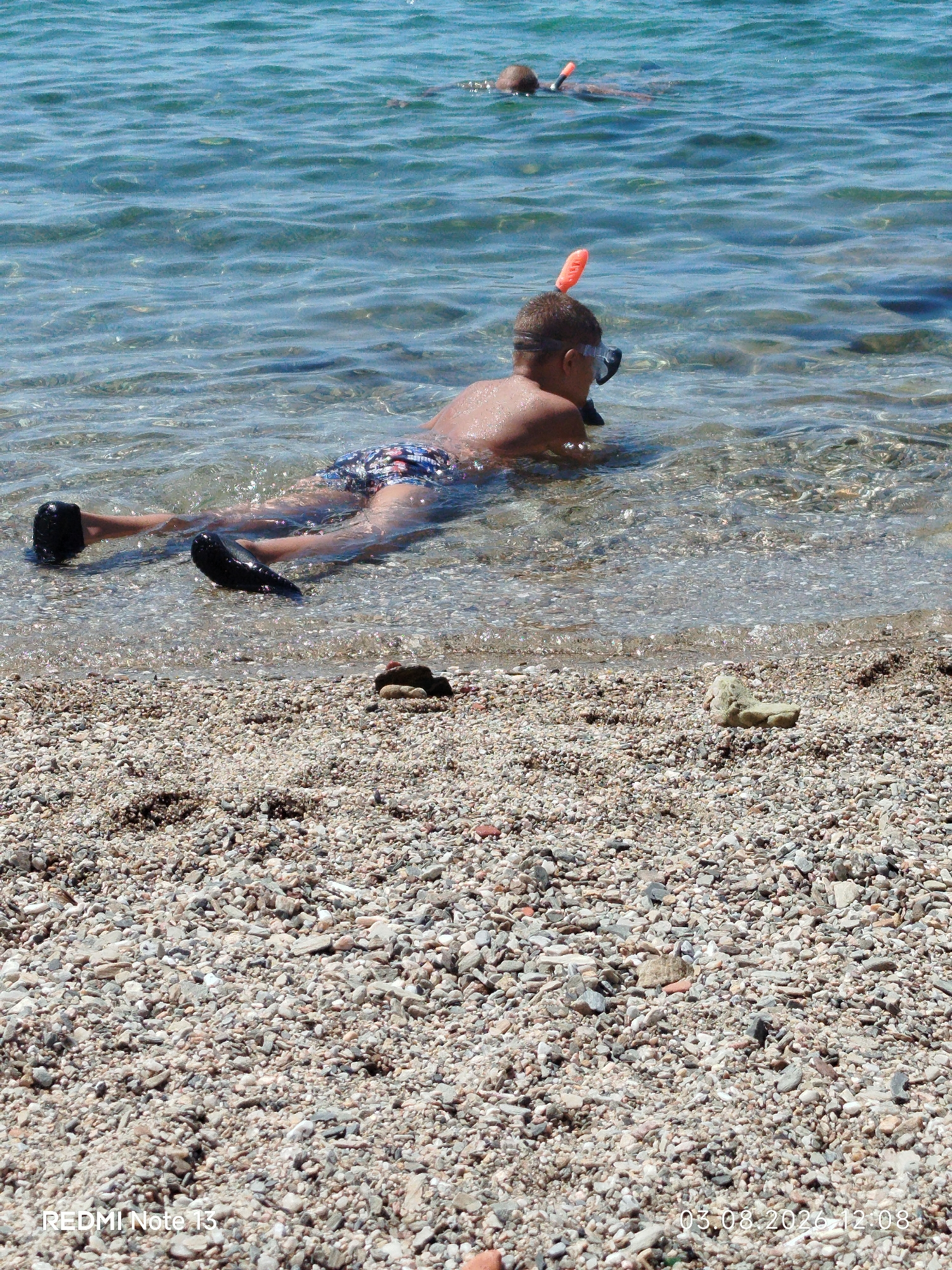 The image depicts a shallow body of clear, blue water lapping onto a pebbled beach. In the foreground, a young boy lies on his stomach in the water, wearing a snorkel mask and an orange snorkel tube. He has on patterned swim trunks and black water shoes. His legs are extended behind him, partially submerged. Further in the water, another person, possibly a child, is also snorkeling, with only their head and snorkel visible. The beach is covered in a variety of small, light-colored pebbles. The water is calm with gentle ripples. The scene appears to be a sunny day at a coastal location in Sithonia Municipal Unit, Greece.