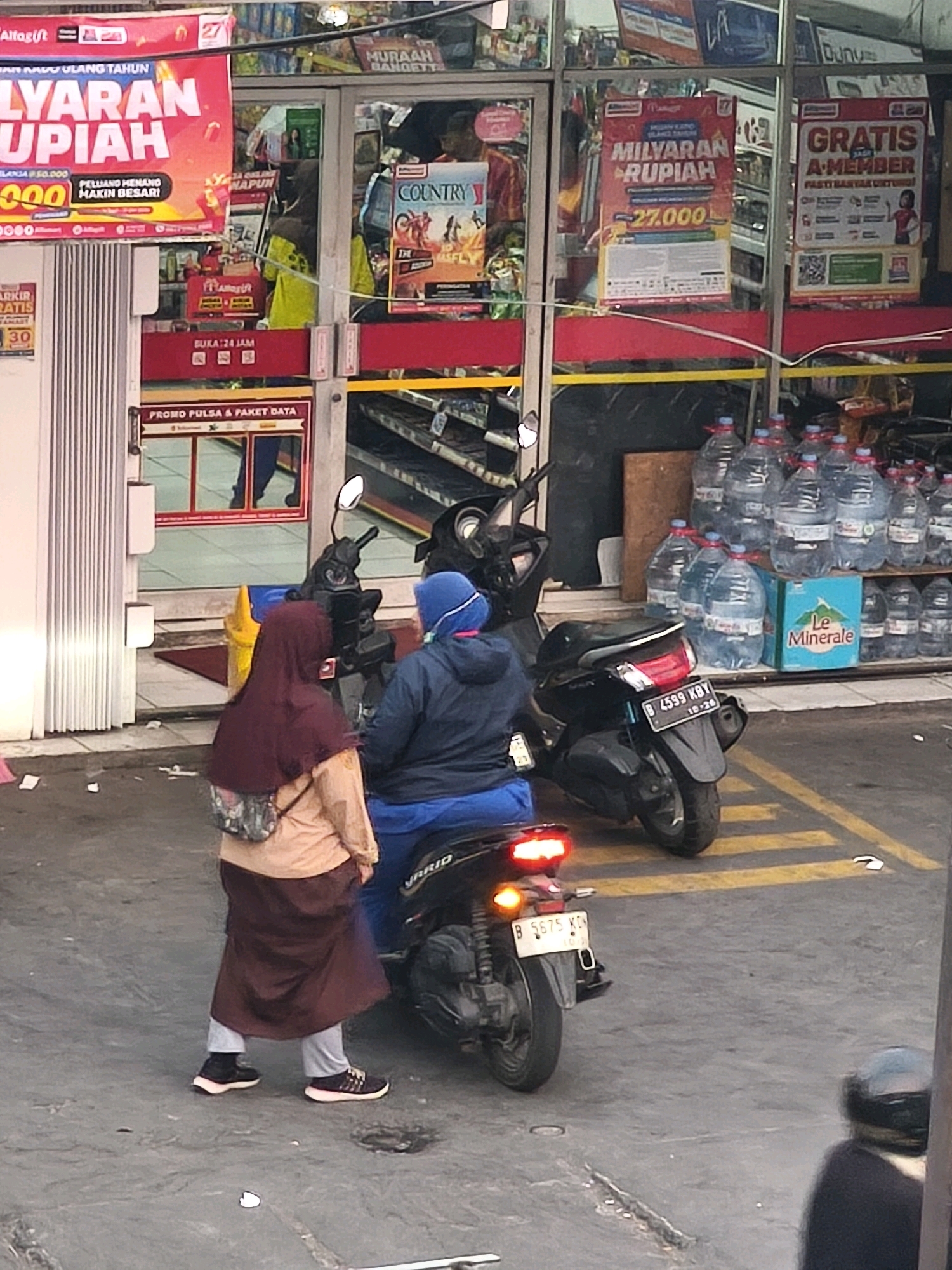 The image depicts the exterior of a minimarket, likely in Bekasi, Indonesia. A red banner advertising a "MILYAARAN RUPIAH" (Billions of Rupiah) promotion hangs above the entrance. To the right, another banner announces "GRATIS A-MEMBER" (Free A-Member), with text below indicating "PASTI BANYAK UNTUNG" (Definitely Many Benefits). Inside the store, shelves are stocked with bottled water, including a prominent display of "Le Minerale" brand water bottles.
In the foreground, two individuals are near parked motorcycles. One person, wearing a maroon hijab and a beige top with a patterned bag, stands facing away from the camera. The other person, wearing a blue jacket and a blue hijab, is seated on a motorcycle, also facing away. A third motorcycle is parked behind them. The ground is asphalt with yellow painted lines. The overall scene suggests a retail environment with promotional activities and common urban elements like parked motorcycles.