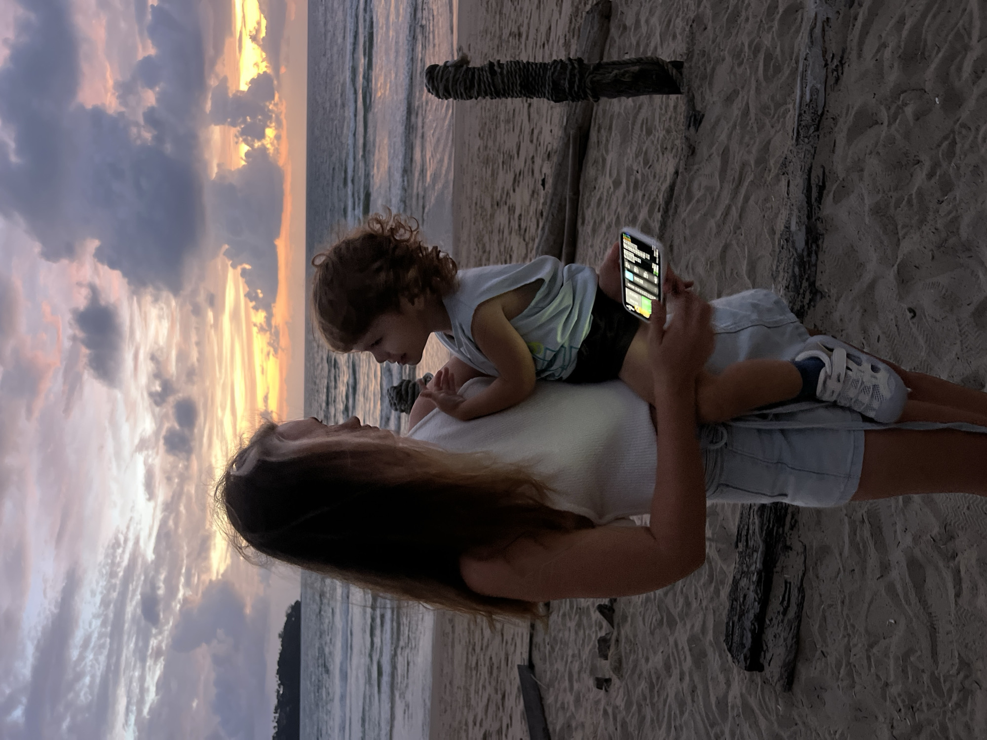In a medium shot, a woman stands on a sandy beach, holding a young child. The woman has long, dark hair and wears a white tank top and light blue denim shorts. The child, with curly brown hair, is facing the woman and wearing a light blue sleeveless shirt and dark shorts. The woman is holding a smartphone in her right hand, which displays a colorful screen. In the background, the ocean meets a cloudy sky painted with the soft hues of a sunset, transitioning from yellow and orange near the horizon to purple and gray higher up. The beach is scattered with pieces of driftwood, and a thick rope is tied around a weathered wooden post to the right of the frame. The scene appears to be located in Choeng Thale, Thailand, given the environmental context of a tropical beach at sunset.