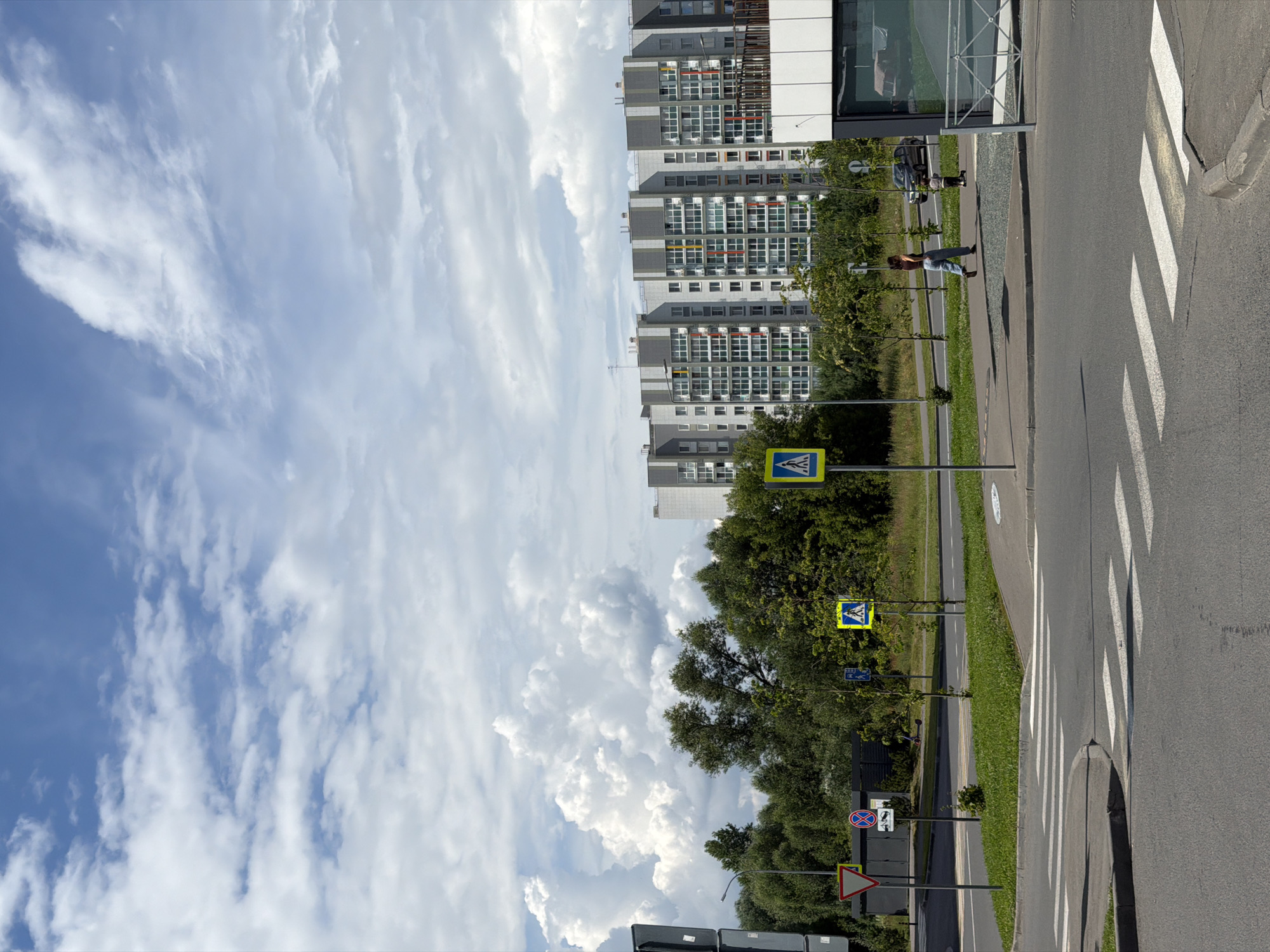 The image depicts a sunny day scene in a modern urban environment, likely in Kazan, Russia. In the foreground, a pedestrian crossing is marked with white stripes on asphalt. To the right, a sidewalk runs parallel to the road, with a woman walking and a child on a scooter. Beyond the sidewalk, a line of green trees forms a boundary, behind which stands a multi-story residential building with a gray facade and numerous windows, accented with vertical stripes of color (red, yellow, and green). The sky is predominantly blue with scattered white, cumulus clouds. Several traffic signs are visible, including a yield sign, a "no stopping" sign, and pedestrian crossing signs. A street lamp is also present. The overall impression is one of a well-maintained, residential area with pedestrian and vehicular traffic.