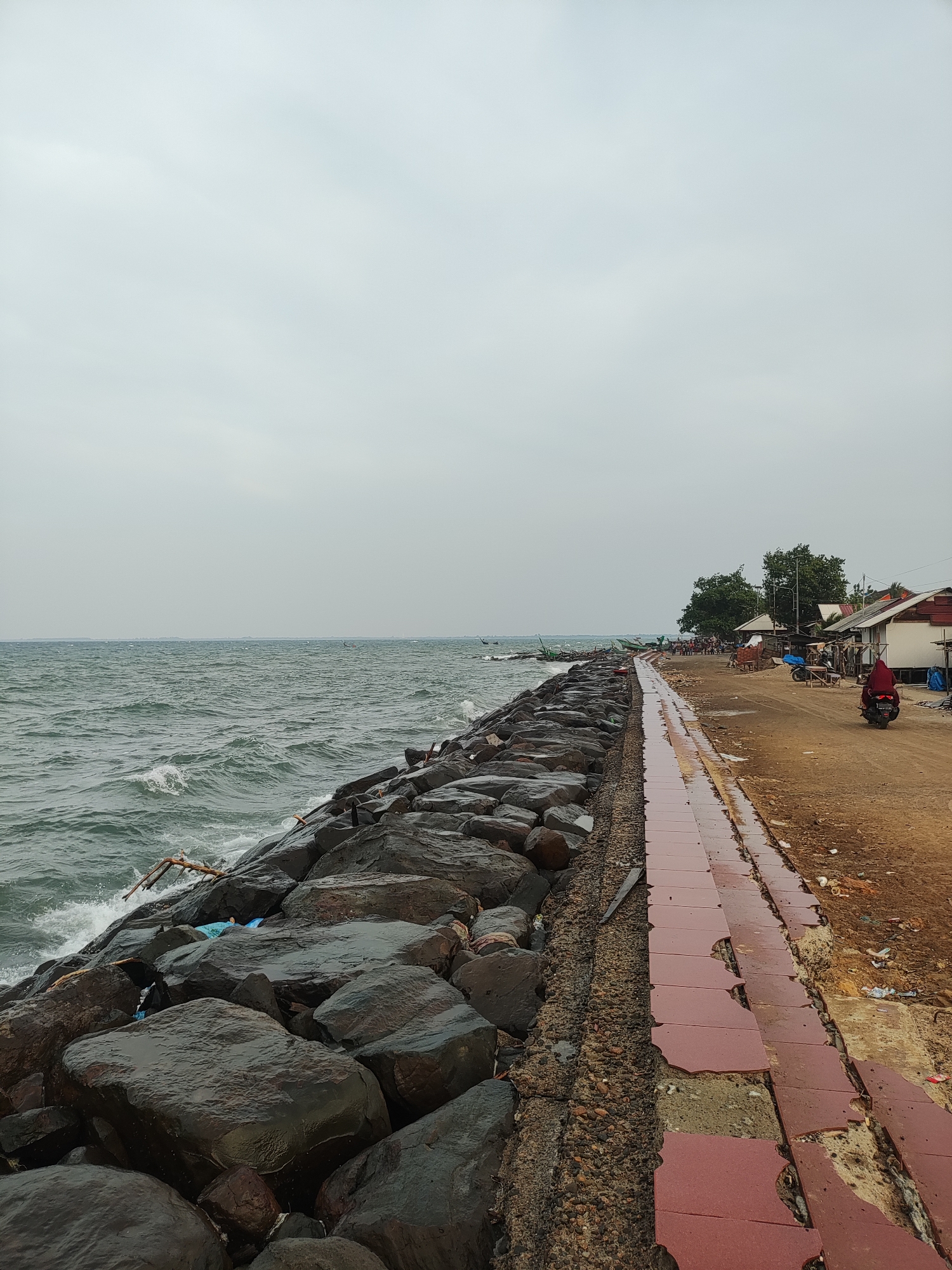 The image shows a coastal scene in Lhokseumawe, Indonesia, under an overcast sky. The shoreline is protected by a rock breakwater, with waves crashing against it. Adjacent to the rocks is a dilapidated walkway paved with red tiles, some of which are broken or missing. Beyond the walkway is a dirt road, and a single person is seen riding a scooter on the dirt road. Further down the road, there appear to be some small buildings, and a few people gathered near some boats. A piece of debris lies among the rocks.